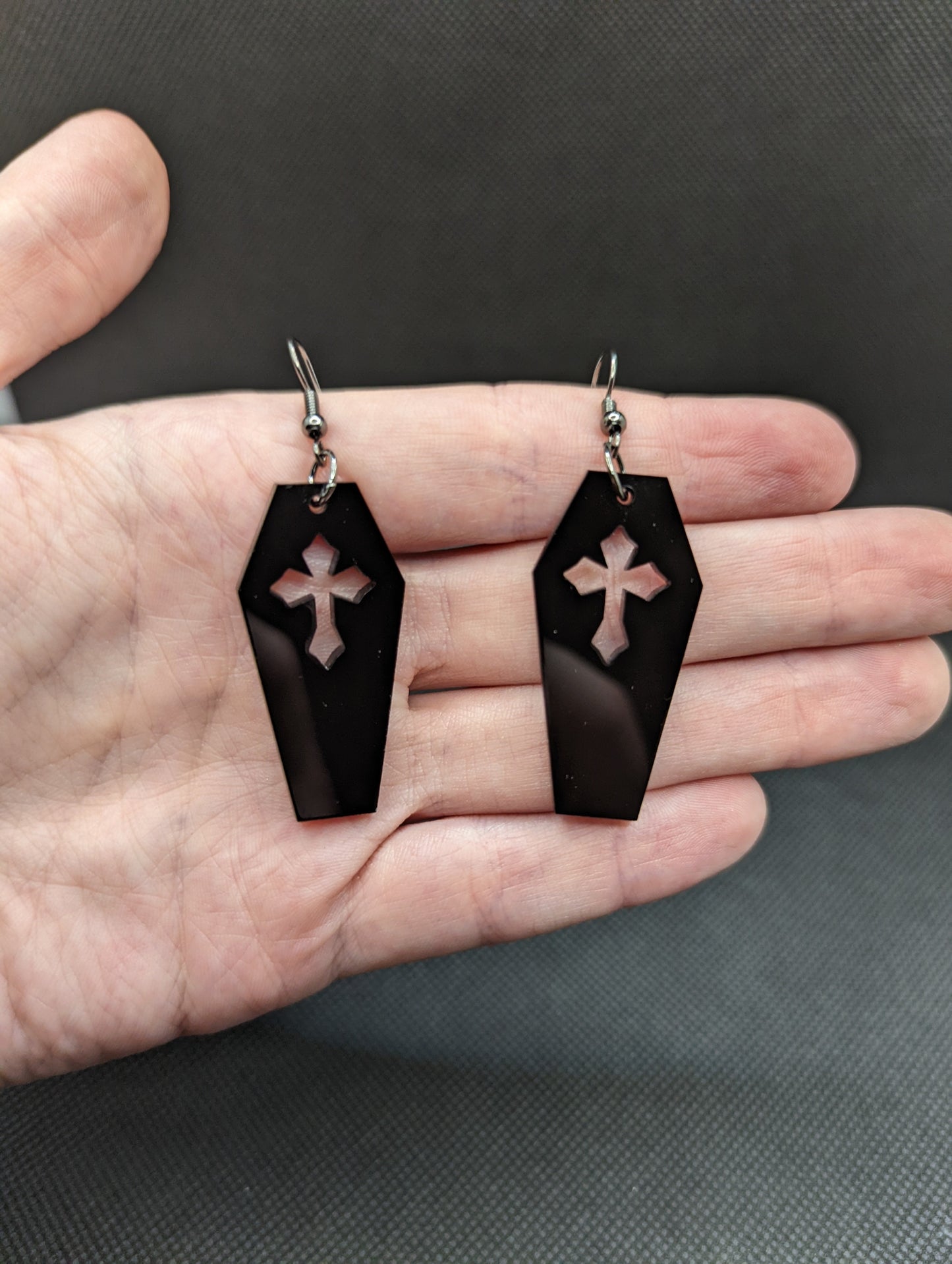 Image of a pair of Halloween coffin / Casket shaped earrings on a hand showing the size against a dark background, they are two and a half finger widths length and one and a half wide. They are black and have silver fixings.