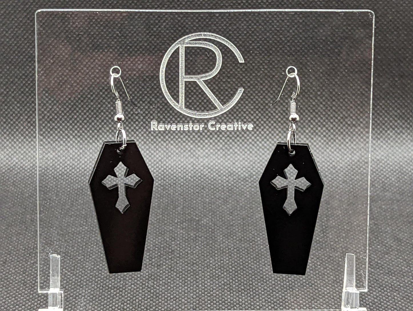 Shows a pair of earrings with silver ear wire hook fixings. They are Spooky Halloween coffins / Casket shapes. The colour of them is black. They are on a see through stand that has the Ravenstor Creative Logo and its against a dark background.
