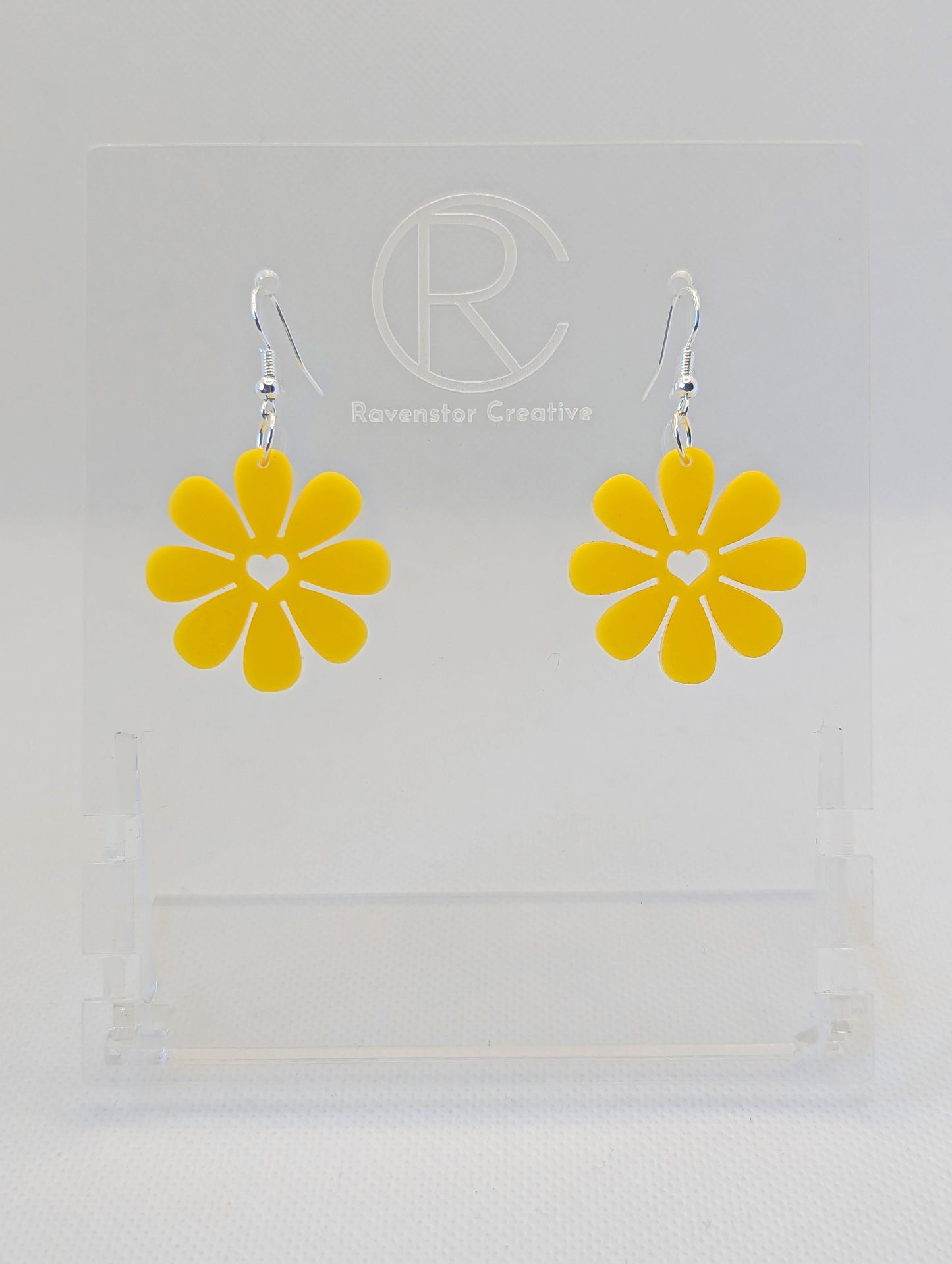 Shows a pair of acrylic earrings that have Silver ear wire hook fixings. They are Daisy Flower earrings with a heart cut out the middle. They are Yellow in colour and are against a white background on see through stand.