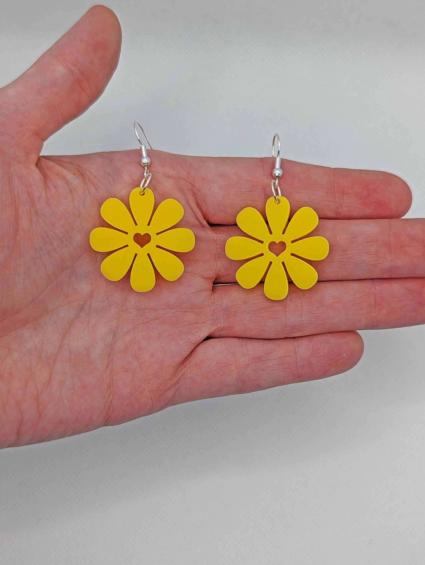 Shows an image of a pair of Daisy flower earrings with a heart cut out the middle. They are on a hand showing the size against a white background, they are two finger widths length and same again wide. They are Yellow and have Silver fixings.