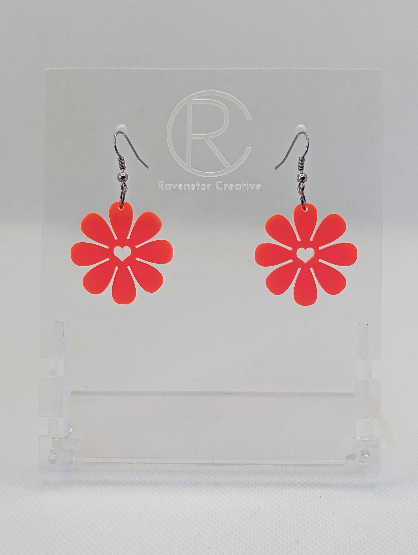 Shows a pair of acrylic earrings that have Black ear wire hook fixings. They are Daisy Flower earrings with a heart cut out the middle. They are Neon Pink in colour and are against a white background on see through stand.