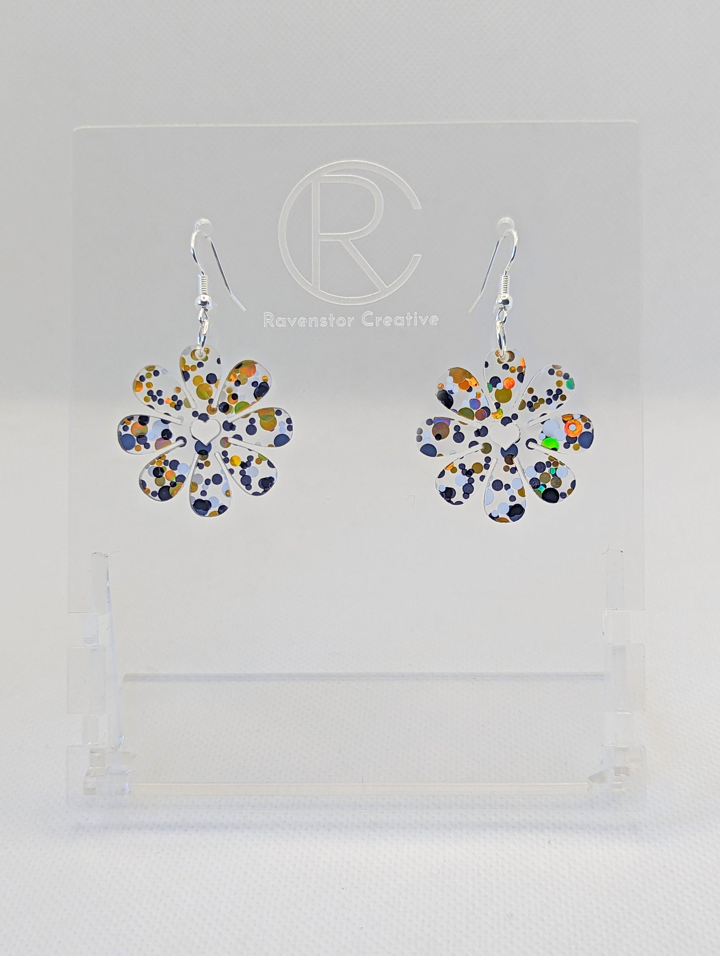 Shows a pair of acrylic earrings that have Silver ear wire hook fixings. They are Daisy Flower earrings with a heart cut out the middle. They are Lollipop Glotter in colour and are against a white background on see through stand.