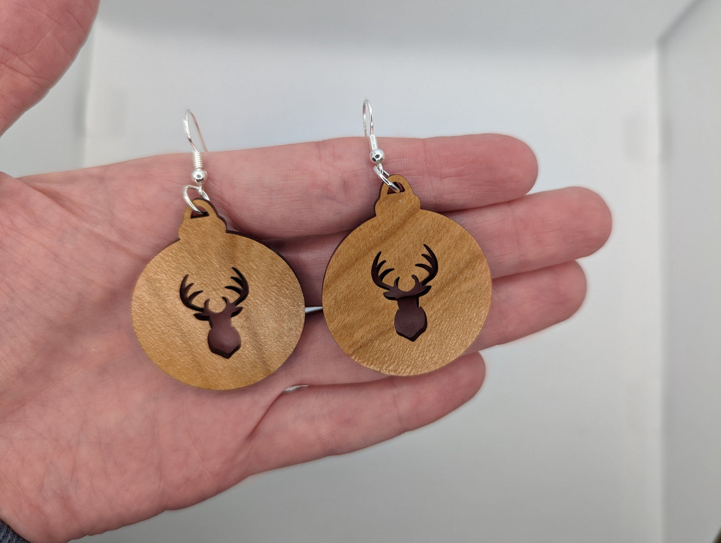 Image of a pair of earrings. They are Christmas / Xmas bauble shapes with a deers head cut out in Cherry Wood on a hand showing the size against a white background, they are 2 finger widths length the same again wide. They and have silver fixings.
