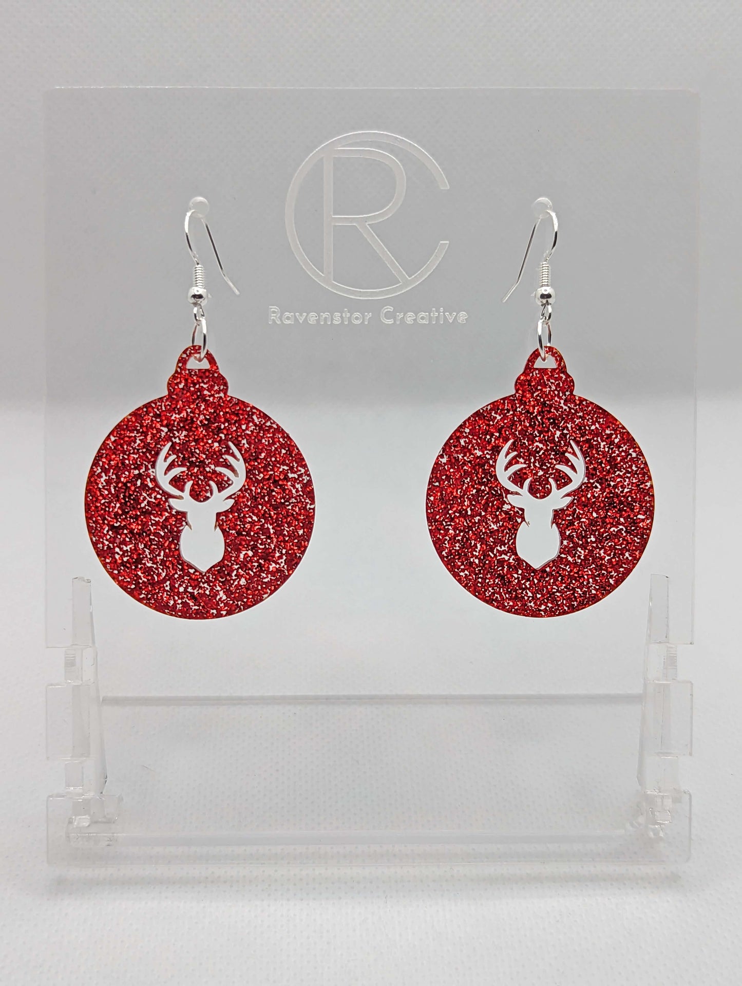 Shows a pair of acrylic earrings that have silver ear wire hook fixings. They are Christmas / Xmas baubles shapes with a deers head shape cut out the middle. The colour is Red Glitter. They are against a white background on see through stands.