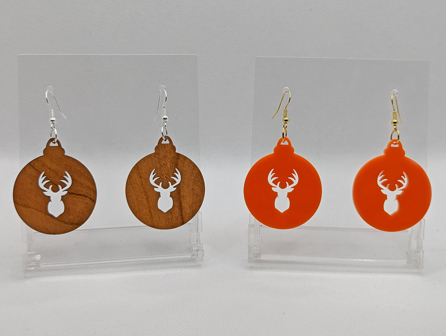 Shows two pairs of earrings that have ear wire hook fixings. They are Christmas baubles with a Deer / Stags head shape cut out the middle. The colours are Cherry Wood and Orange. They are against a white background on see through stands.
