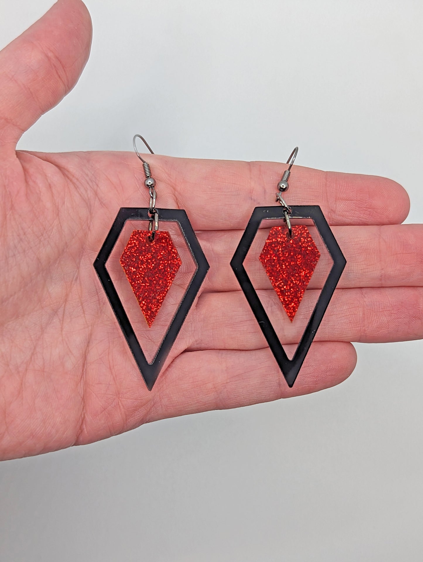 A pair of earrings that are two parts. Black ear wire fixings are attached to a diamond shaped frame with a smaller diamond dangling in the middle. They are on a hand showing the size of three finger widths in length and two wide.