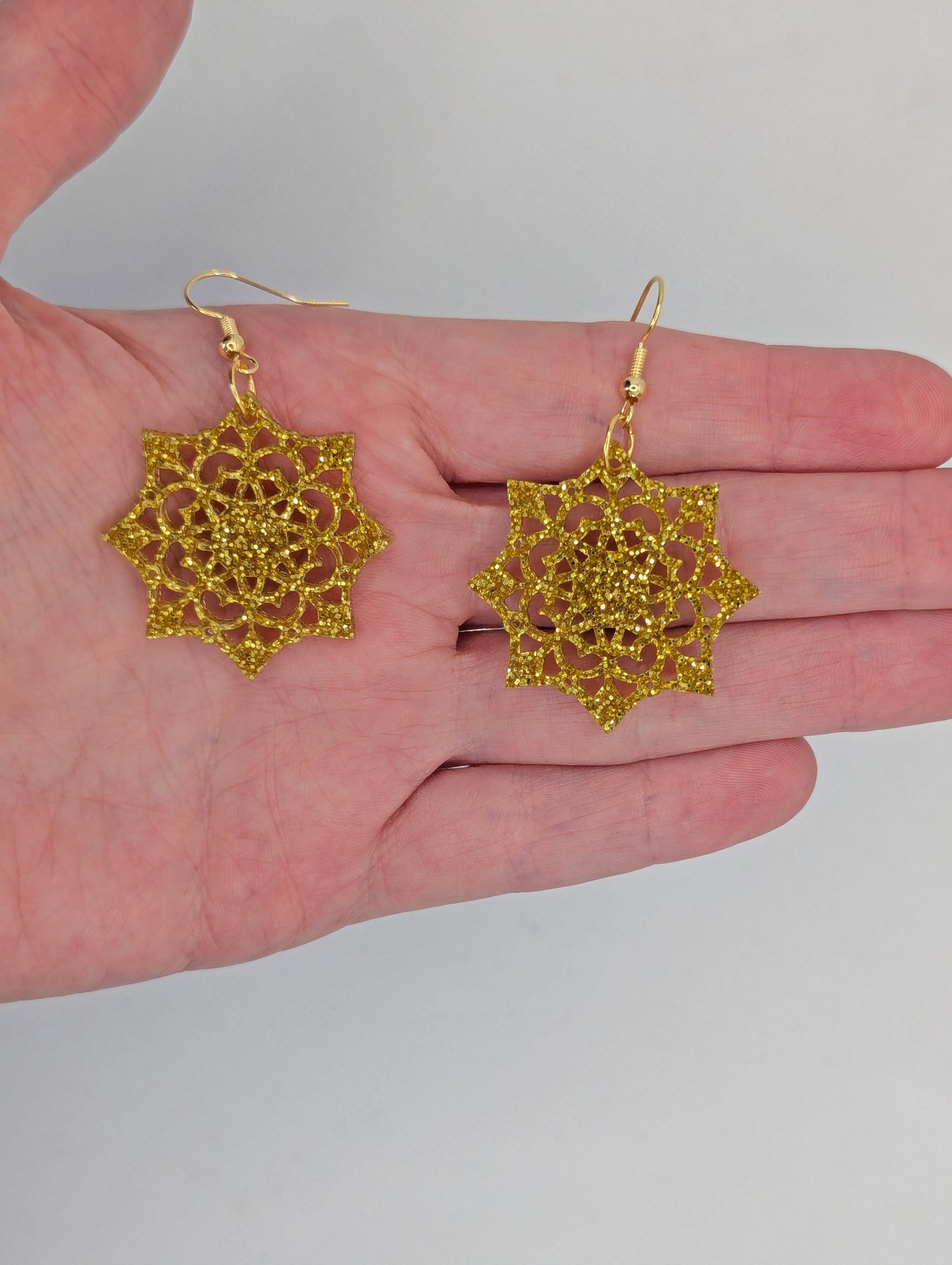 Image of a pair of acrylic earrings with Gold ear wire hook fixings. They are an intricate Mandela pattern with diamond shapes. They are against a hand to show the size of two finger widths long and the same again wide.
