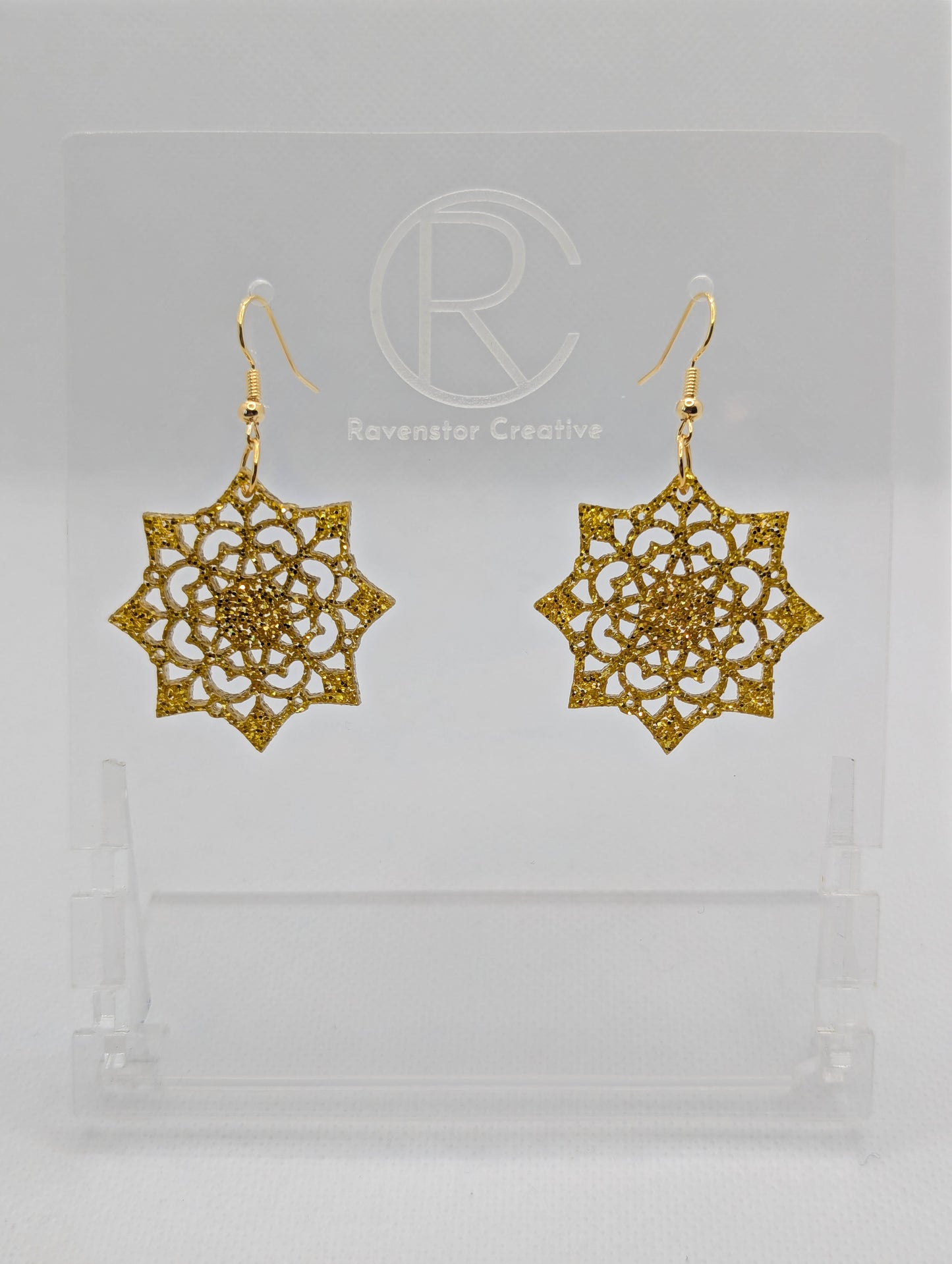 Image of a pair of acrylic earrings with Gold ear wire hook fixings. They are an intricate Mandela pattern with diamond shapes. The colour is Gold Glitter. They are on a clear stand with the Ravenstor Creative logo against a white background.