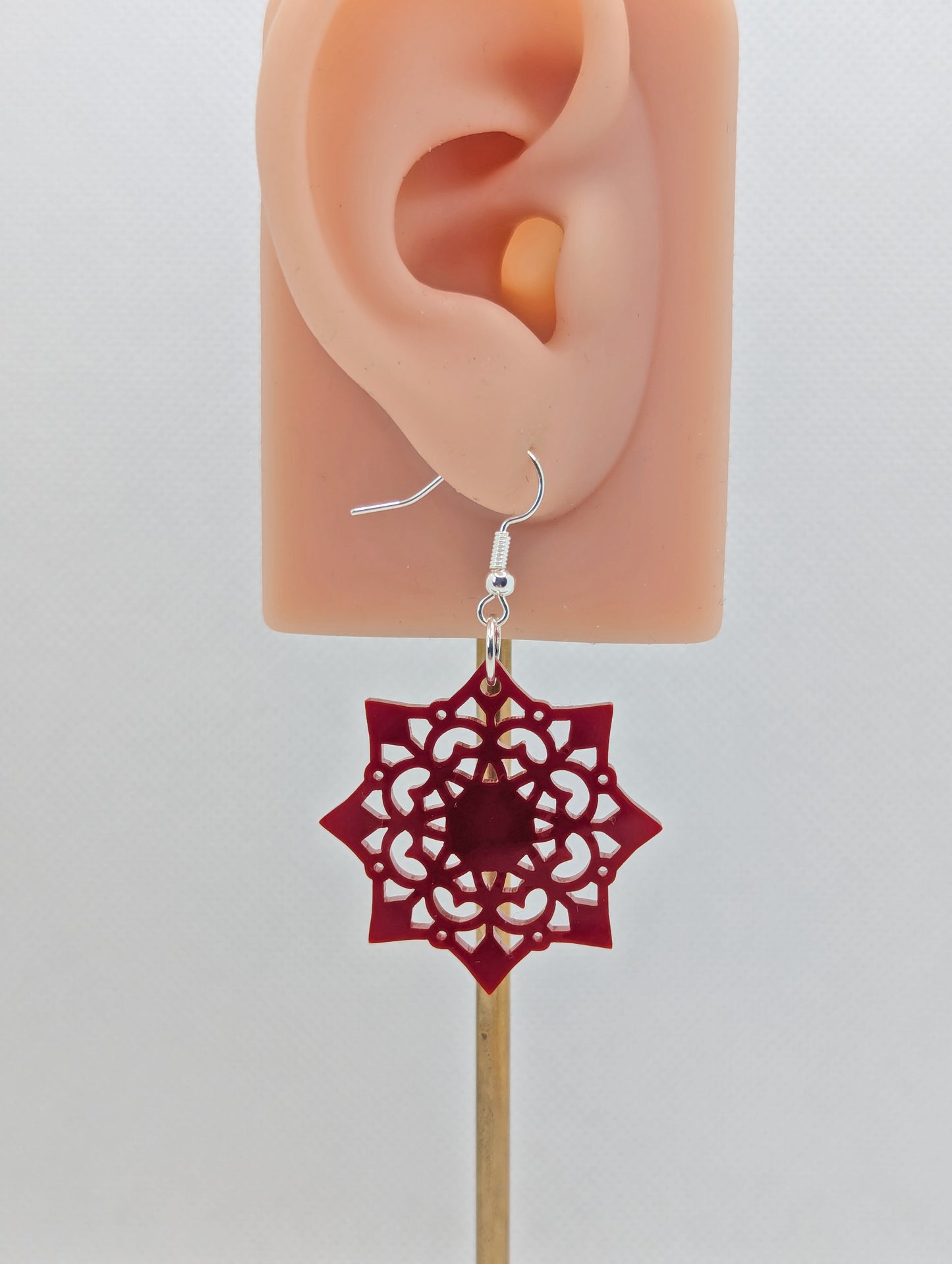 Image of an acrylic earring with a silver ear wire hook fixing. It is an intricate Mandela pattern with diamond shapes. The colour is Maroon. It is on an ear to show the size against a white background.