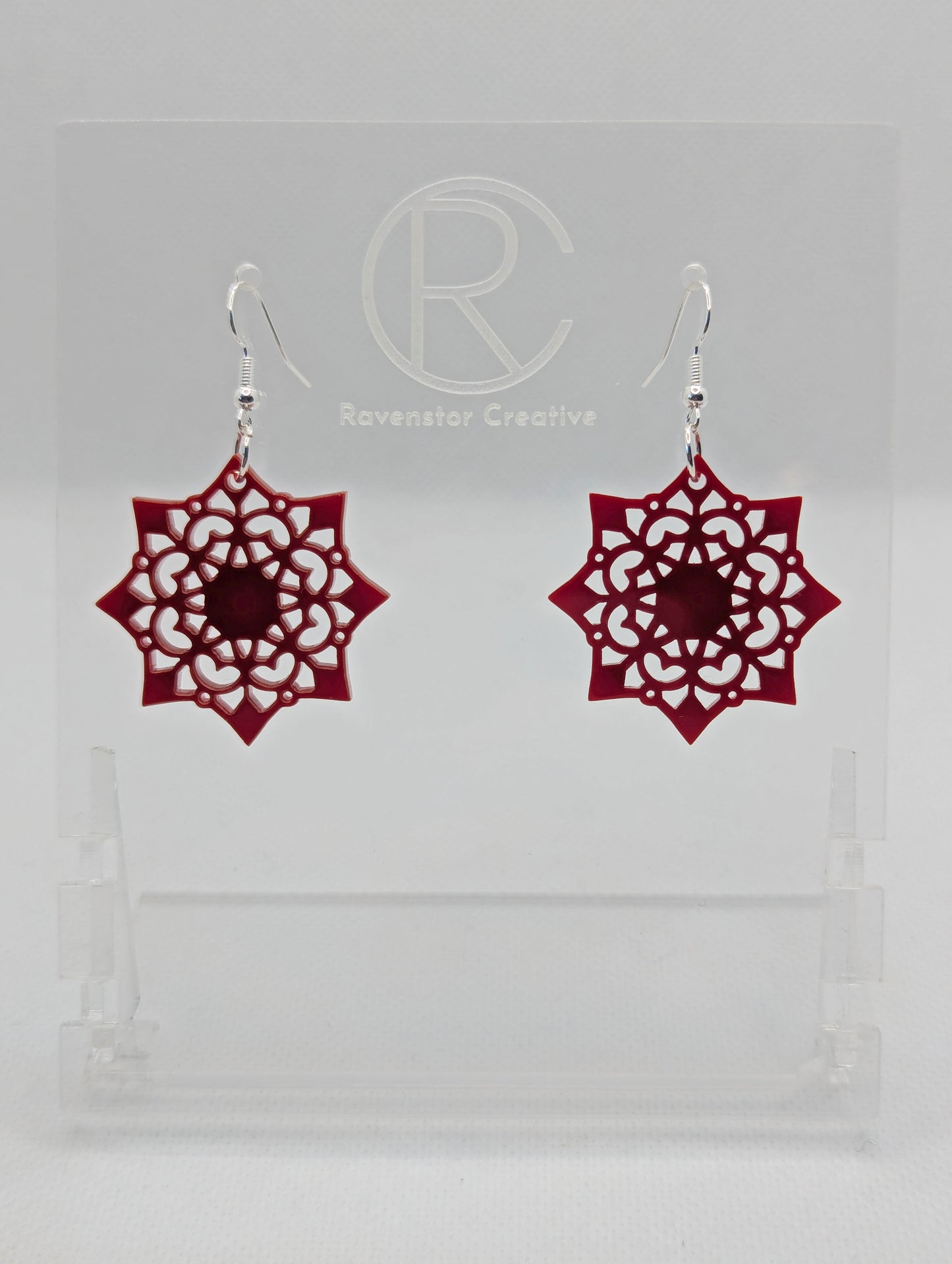 Image of a pair of acrylic earrings with silver ear wire hook fixings. They are an intricate Mandela pattern with diamond shapes. The colour is Maroon. They are on a clear stand with the Ravenstor Creative logo against a white background.