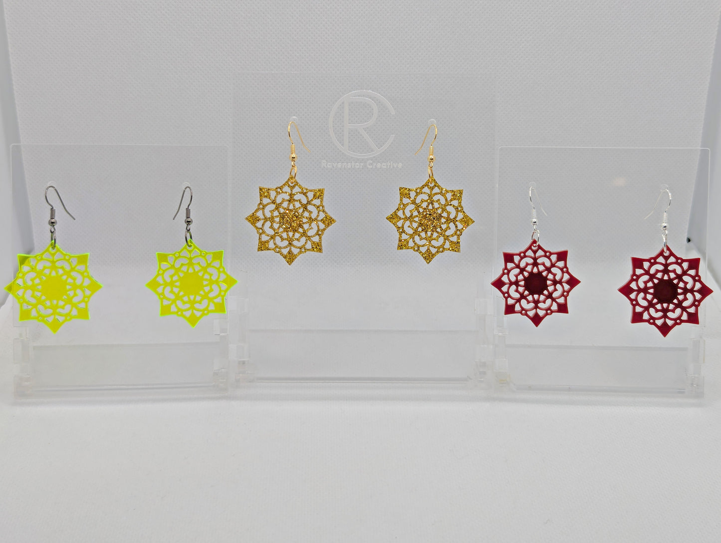 Image of Three pairs of earrings with ear wire hook fixings. They are an intricate Mandela pattern with diamond shapes. The colours are Neon Yellow, Gold Glitter and Maroon. They are on clear stands against a white background.