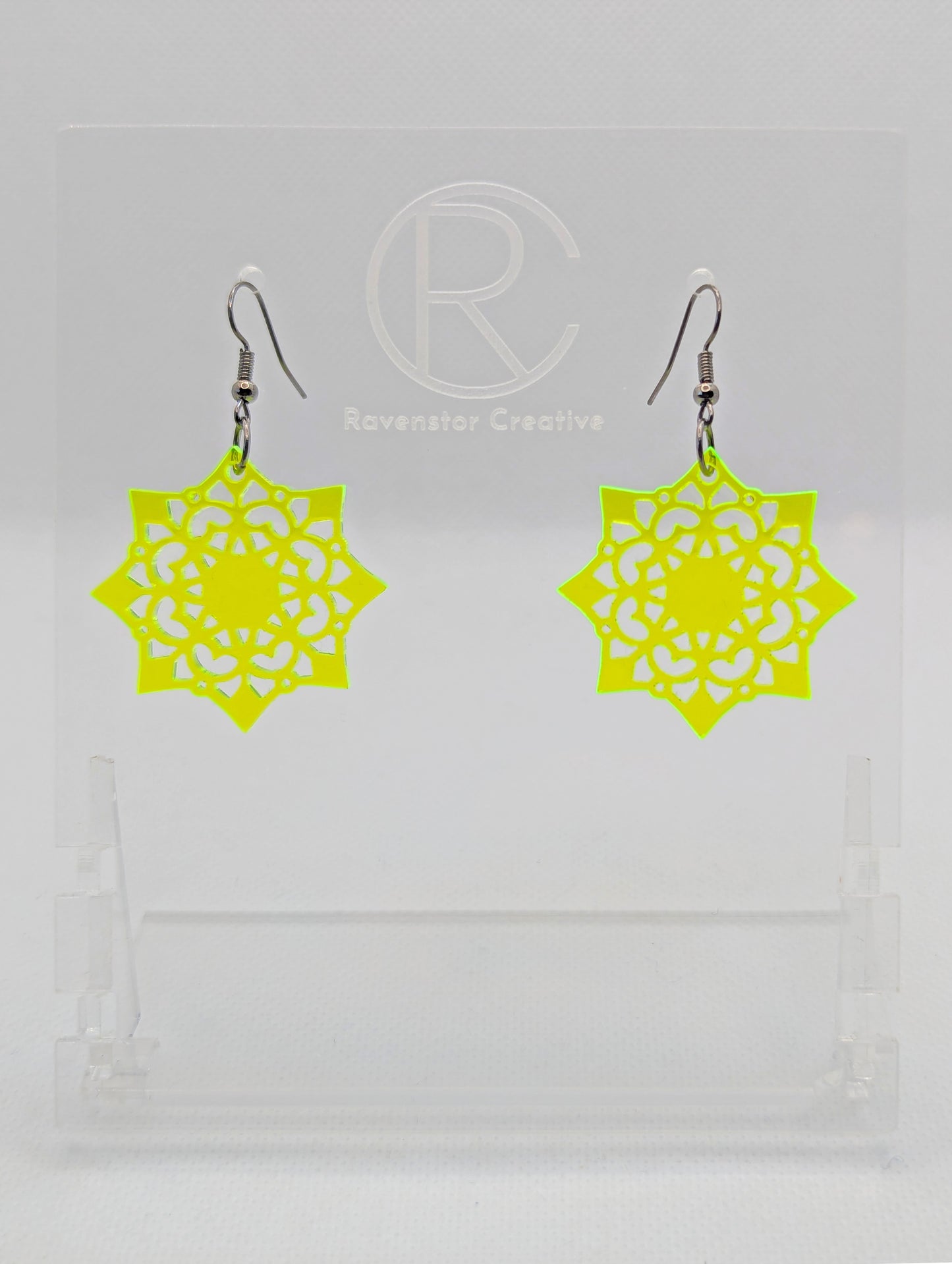 Image of a pair of acrylic earrings with black ear wire hook fixings. They are an intricate Mandela pattern with diamond shapes. The colour is Neon Yellow. They are on a clear stand with the Ravenstor Creative logo against a white background.
