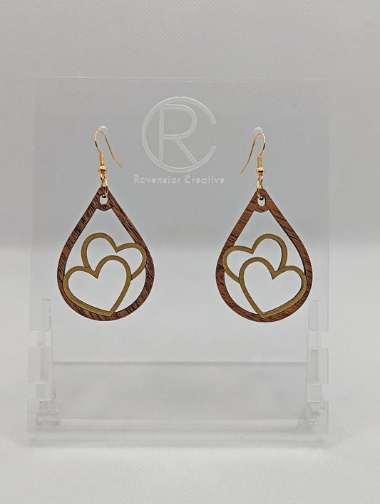 Shows a pair of earrings that have gold ear wire hook fixings. They are teardrop shapes with overlapping hearts in the middle that are painted Gold. The are made from Walnut Wood and are against a white background on see through stand.