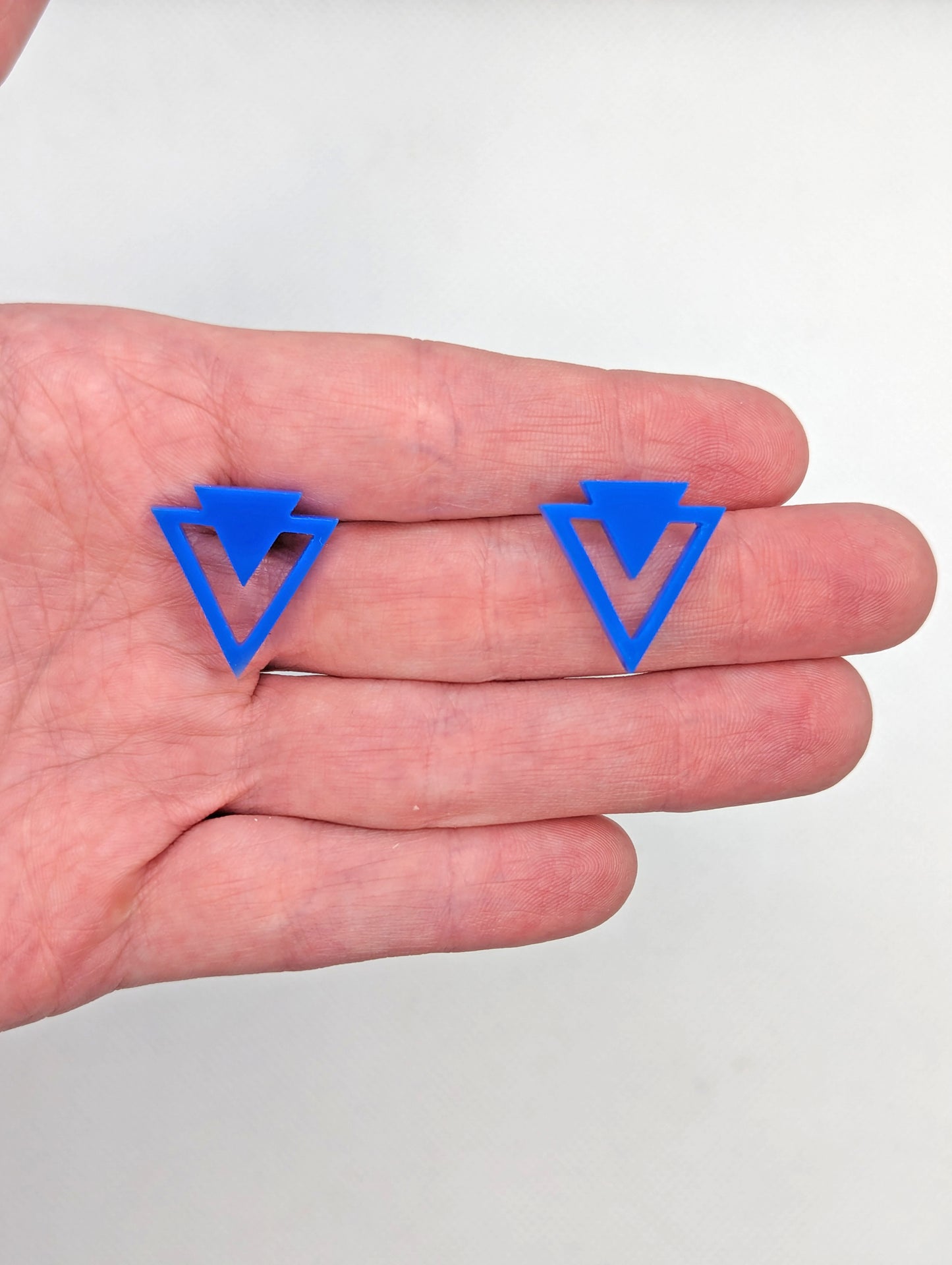 Shows a pair of small studded acrylic earrings that are a triangle with another triangle around it joining at the top. The colour is Blue. They are against a hand showing the size of just over one finger width in length and width.