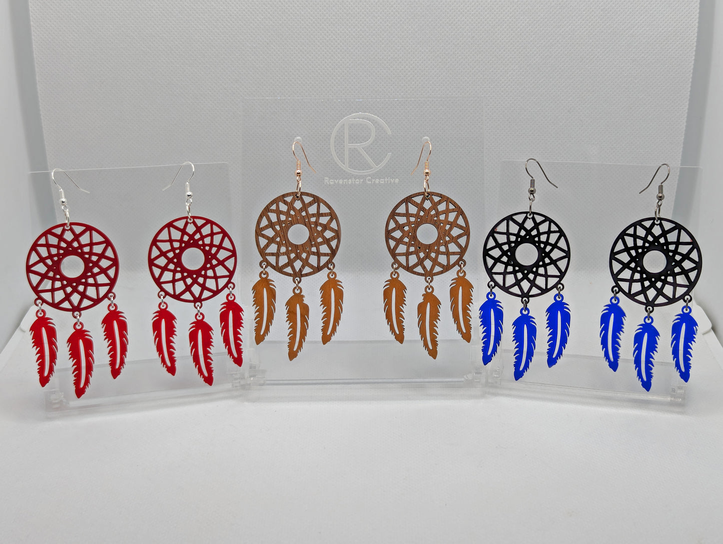 Three pairs of earrings shaped like dreamcatchers made in two parts. A patterned circle at the top with 3 dangling shapes under. the colours are maroon for the top and red for the dangles, walnut wood for top and cherry wood for the bottom, then black for the top and translucent Blue for the bottom
