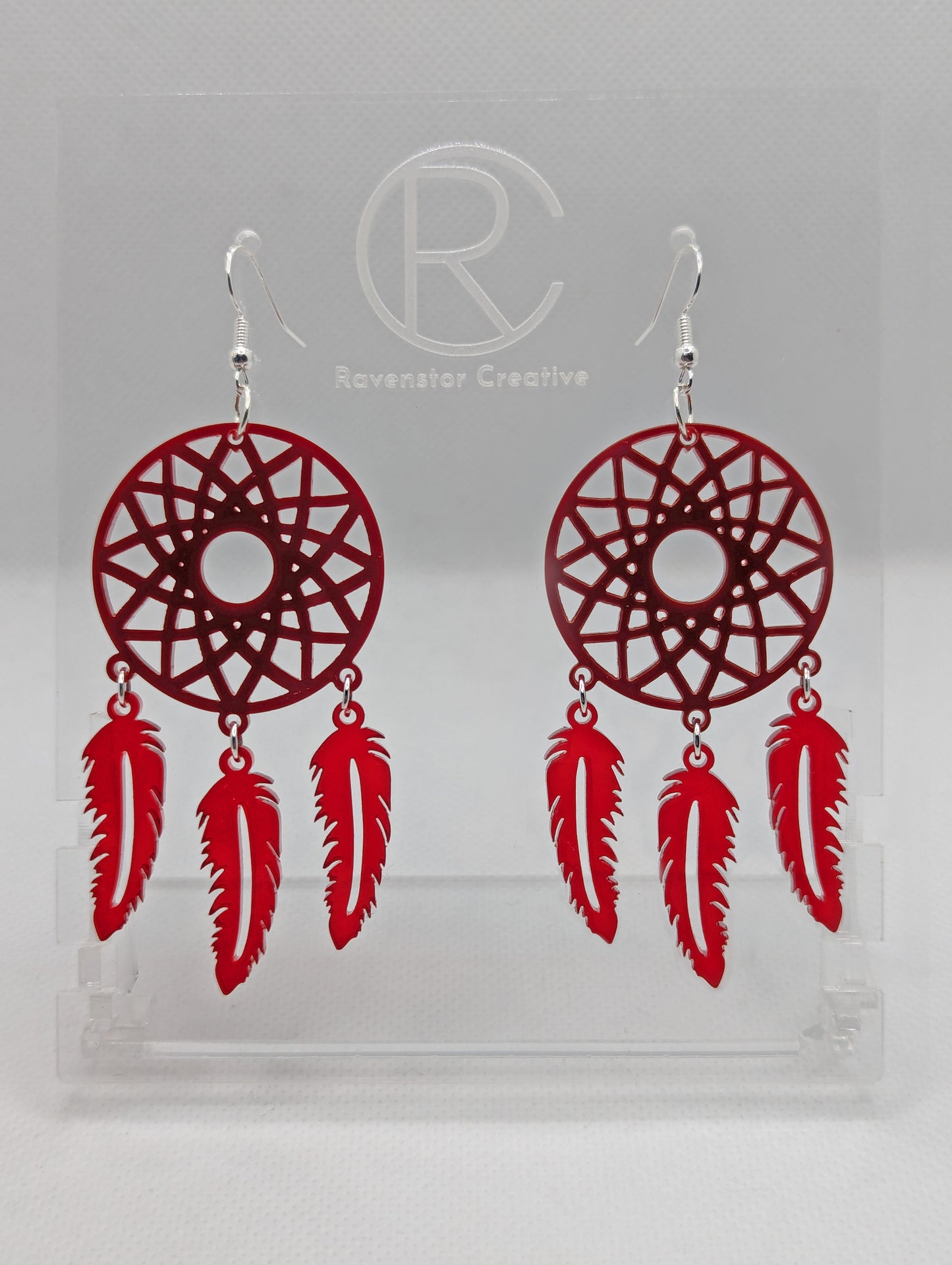 A pair of large earrings shaped like a dreamcatcher. A patterned circle at the top with 3 dangling shapes under. They are made from acrylic and are maroon for the circle and translucent red for the dangles. They have silver ear wire hook fixings.