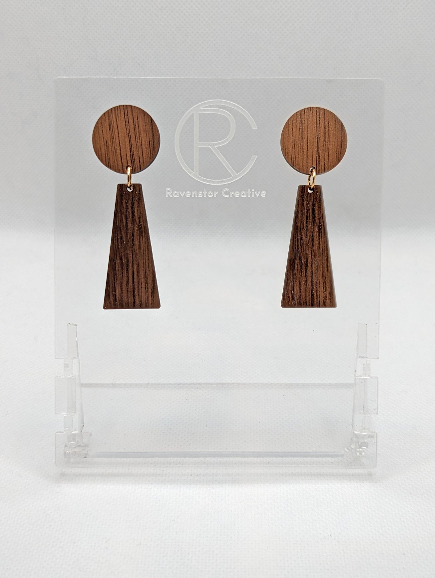 Shows a pair of earrings that are two parts. Stud fixings are attached to an acrylic circle, dangling under is a trapezoid. The colours are cherry wood for the circle and walnut wood trapezoid dangling underneath.