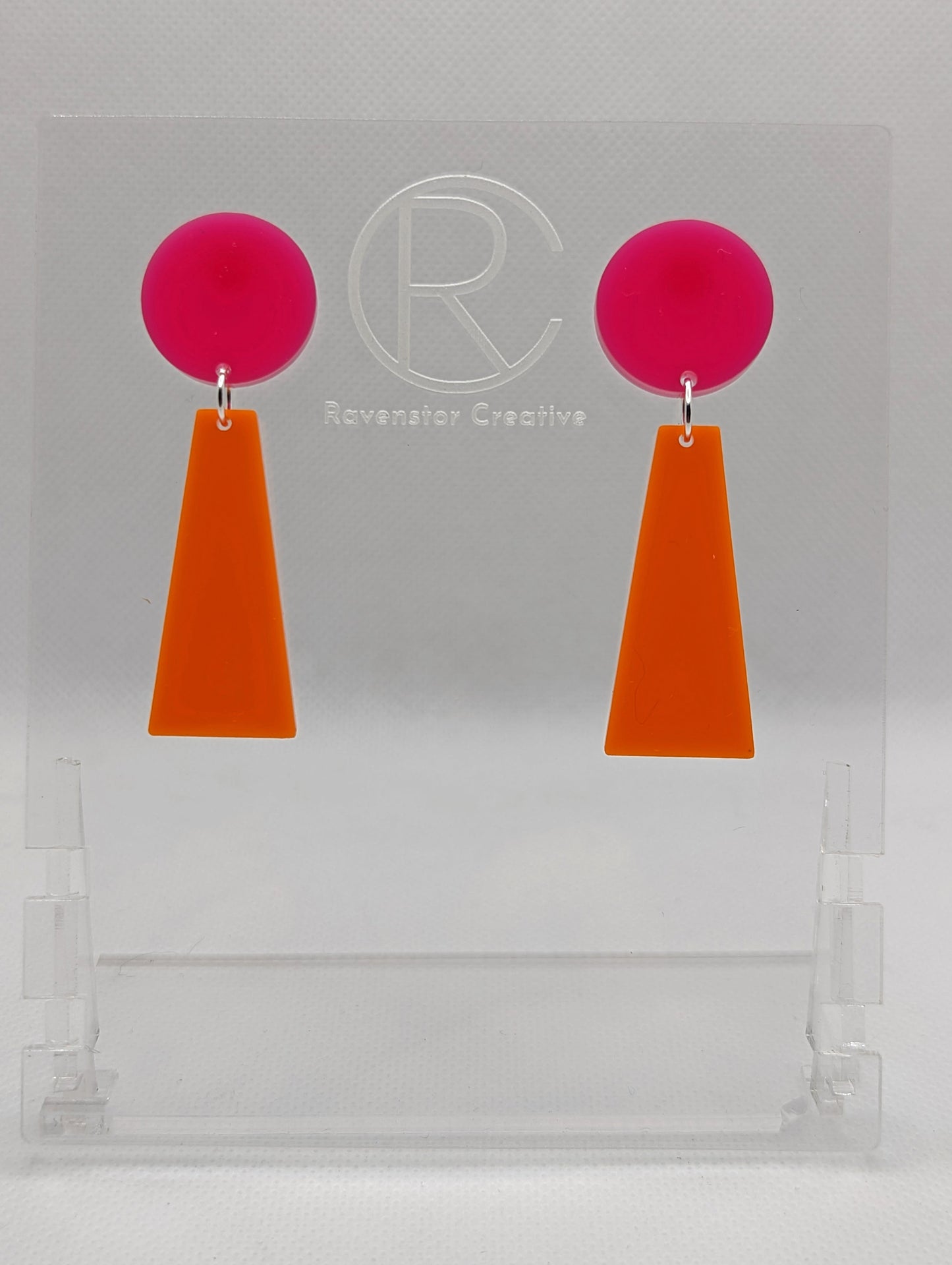 Shows a pair of earrings that are two parts. Stud fixings are attached to an acrylic circle, dangling under is a trapezoid. The colours are hot pink for the circle and grape trapezoid dangling underneath.