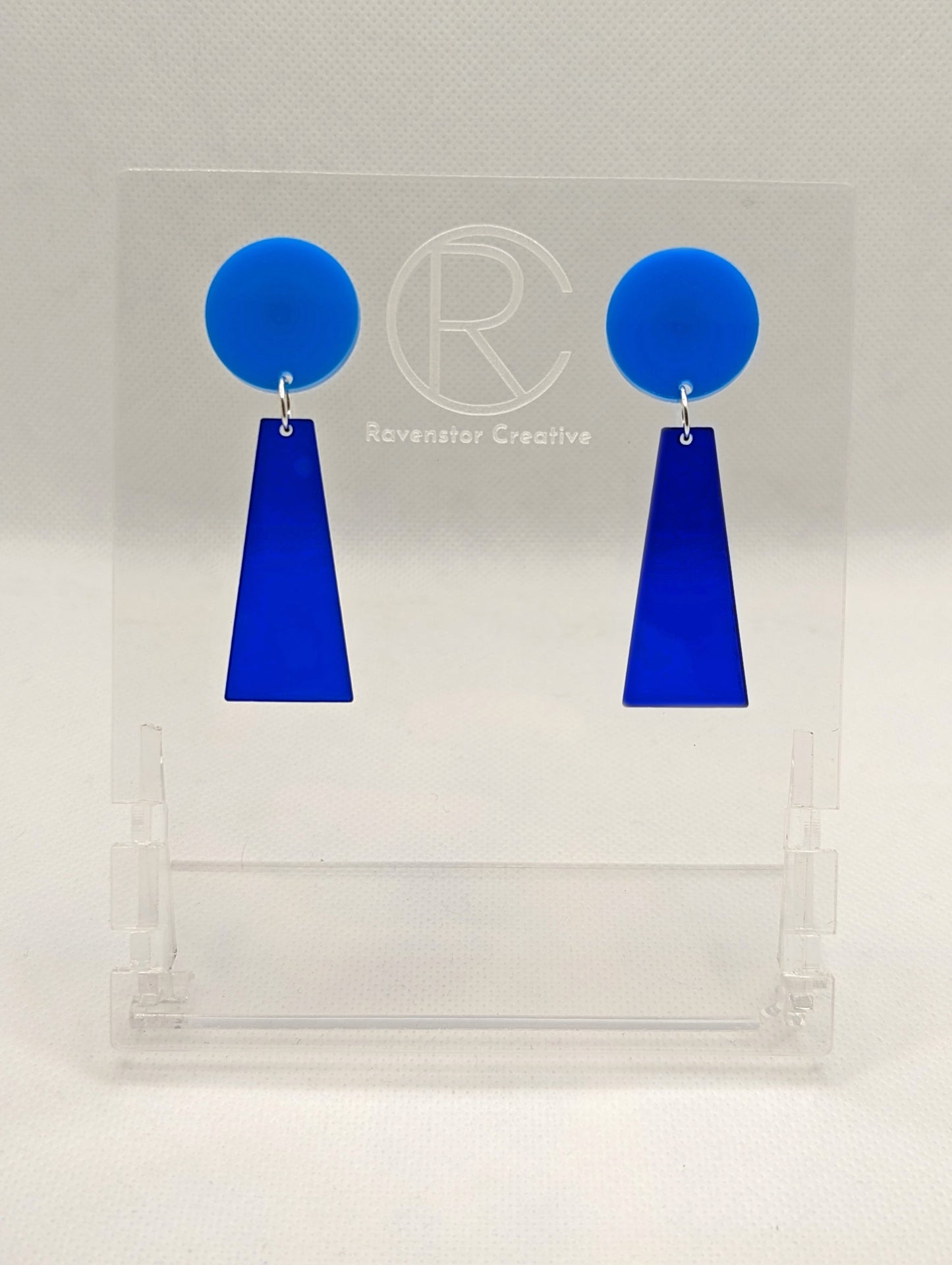 Shows a pair of earrings that are two parts. Stud fixings are attached to an acrylic circle, dangling under is a trapezoid. The colours are blue for the circle and translucent blue trapezoid dangling underneath.