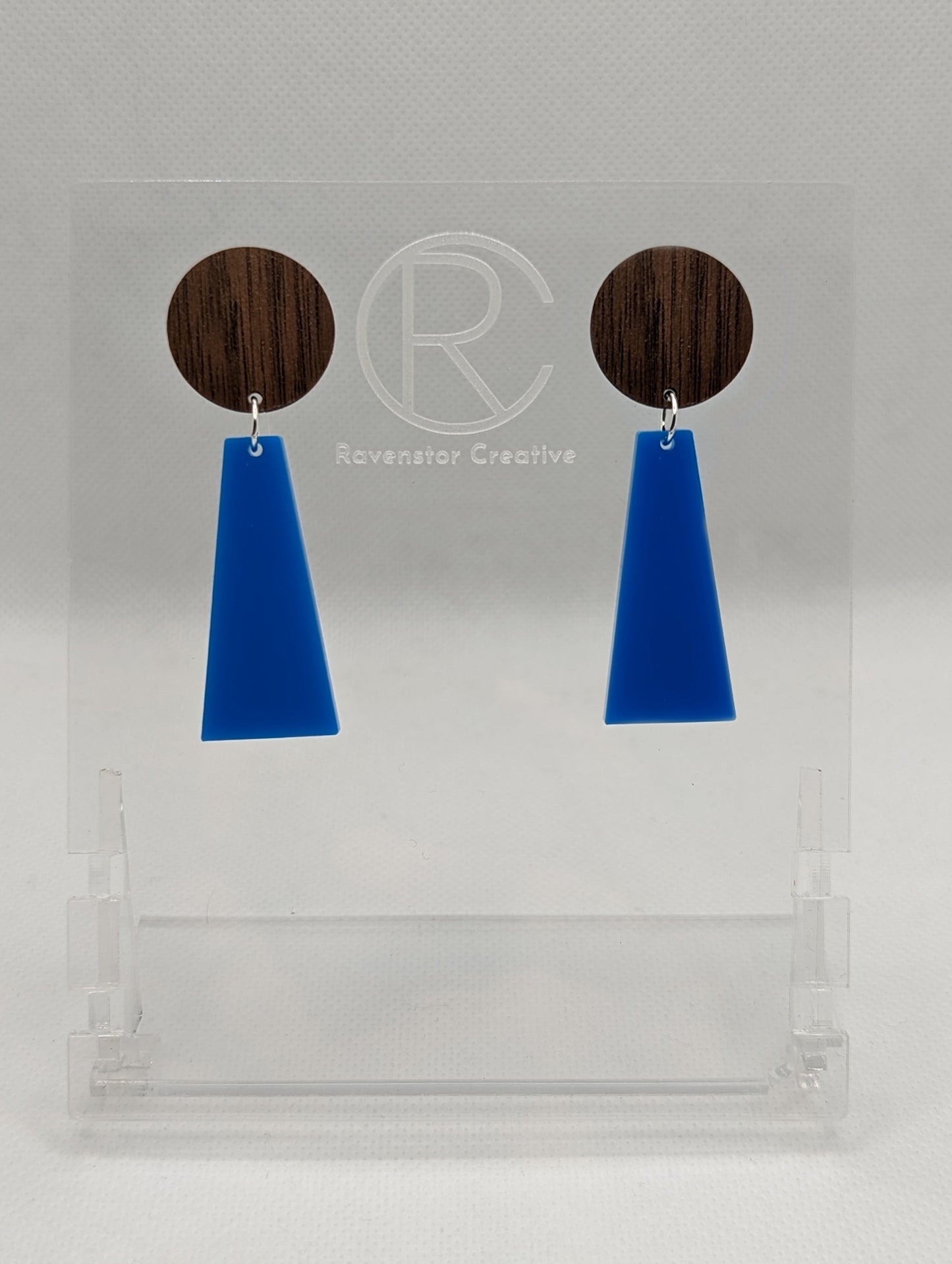 Shows a pair of earrings that are two parts. Stud fixings are attached to an acrylic circle, dangling under is a trapezoid. The colours are walnut wood for the circle and blue trapezoid dangling underneath.