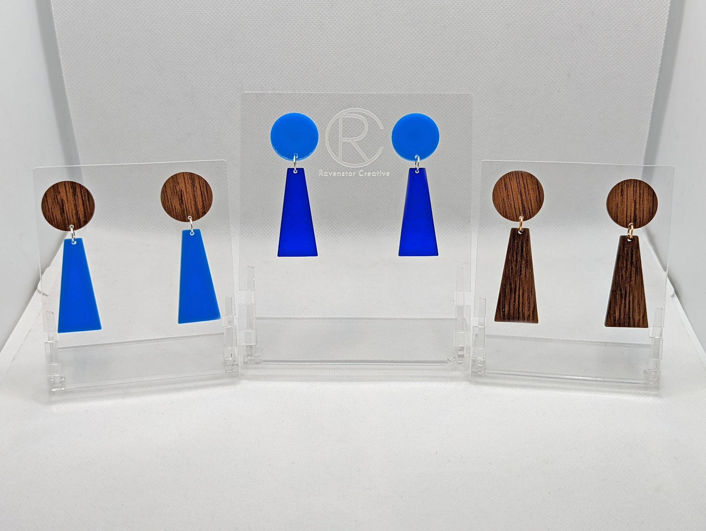 Shows 3 pairs of earrings that are two parts. Stud fixings are attached to an acrylic circle, dangling under is a trapezoid. The colours are walnut wood for the circle and blue for the trapezoid, blue and translucent blue and two woods for both.
