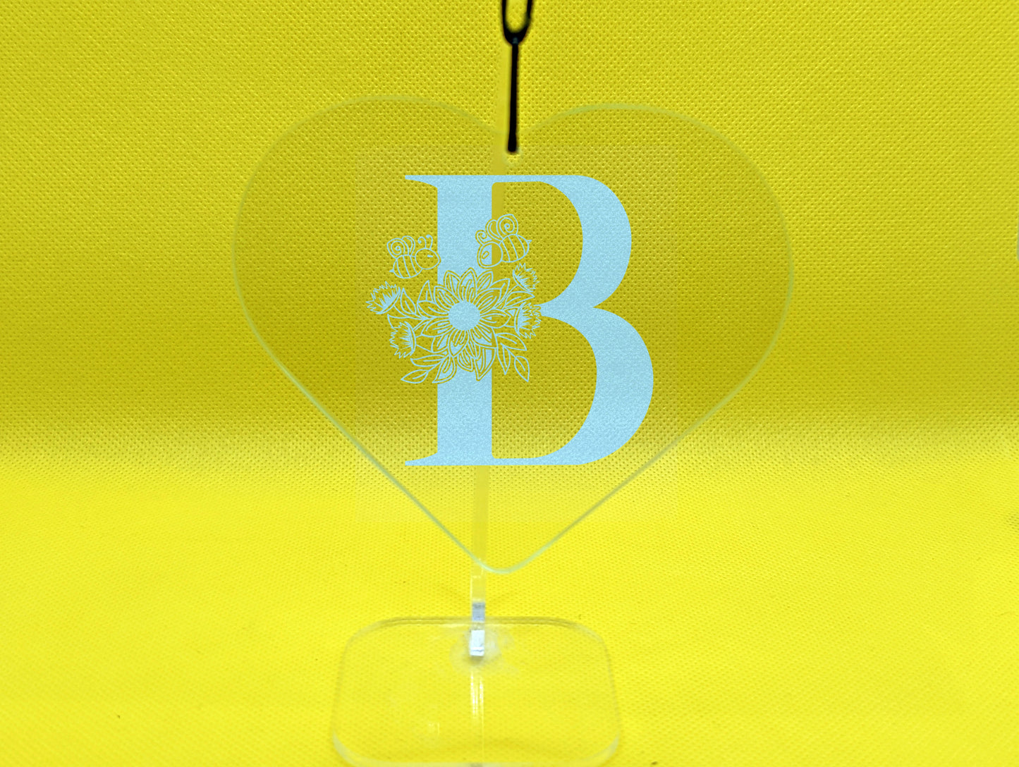 Shows a floral themed hanging glass heart engraved with the example initial B that has a sunflower, leaves and bees on and around it. It is on a clear stand against a yellow background.