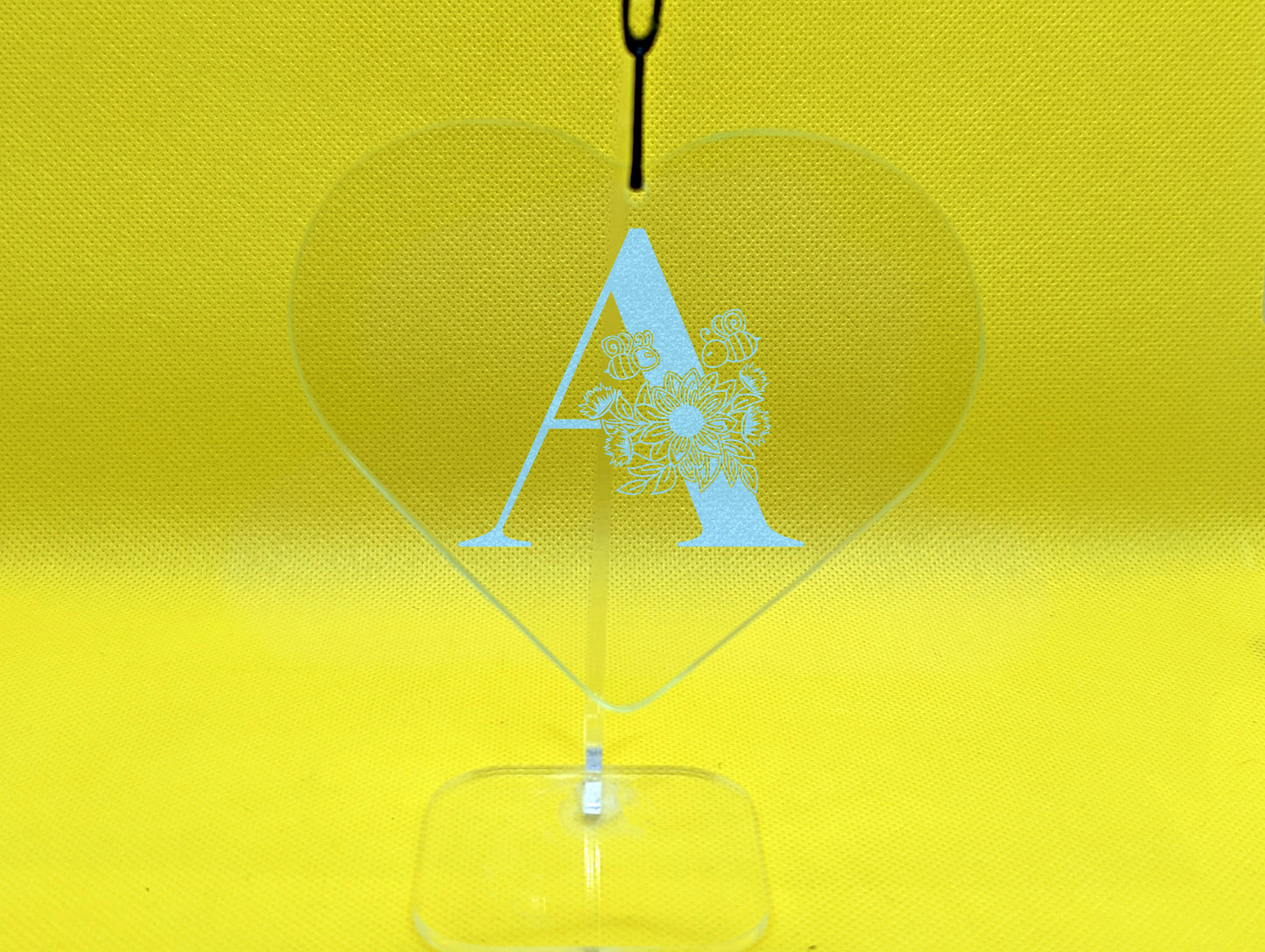 Shows a floral themed hanging glass heart engraved with the example initial A that has a sunflower, leaves and bees on and around it. It is on a clear stand against a yellow background.