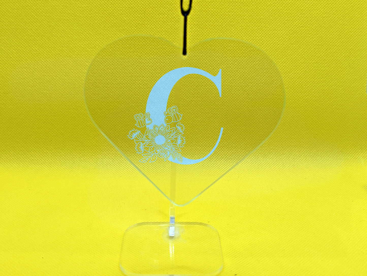 Shows a floral themed hanging glass heart engraved with the example initial C that has a sunflower, leaves and bees on and around it. It is on a clear stand against a yellow background.