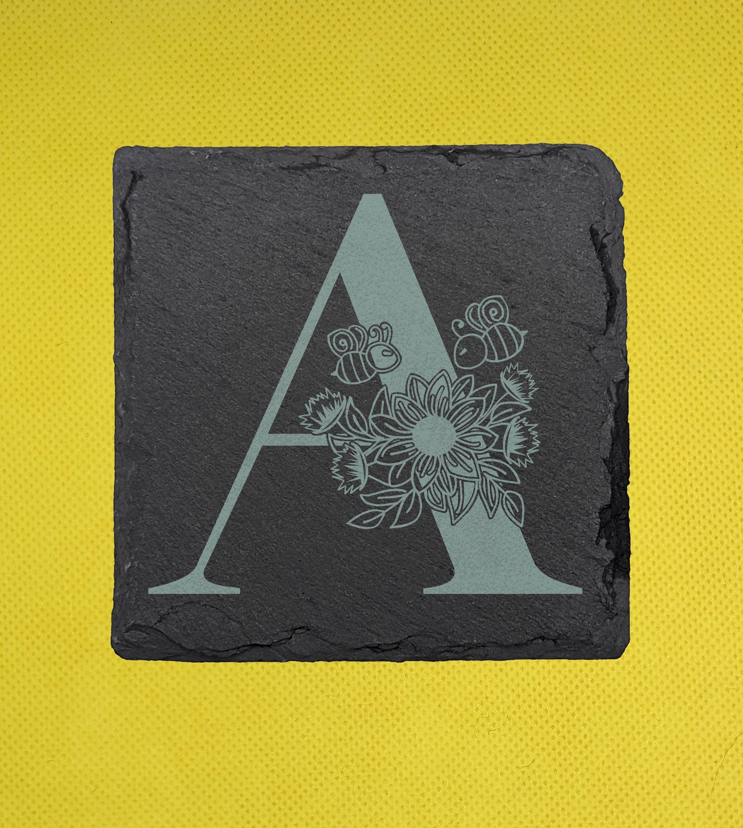 Shows a floral themed square slate coaster engraved with the example initial A that has a sunflower, leaves and bees on and around it. It is on against a yellow background.