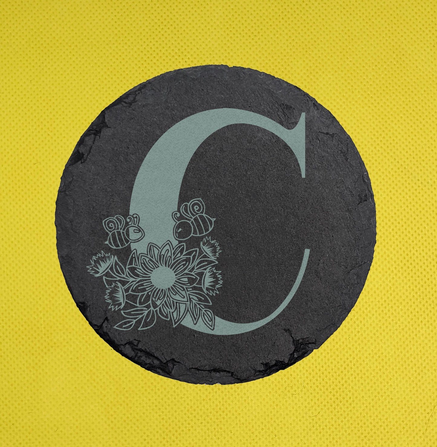 Shows a floral themed circle slate coaster engraved with the example initial C that has a sunflower, leaves and bees on and around it. It is against a yellow background.