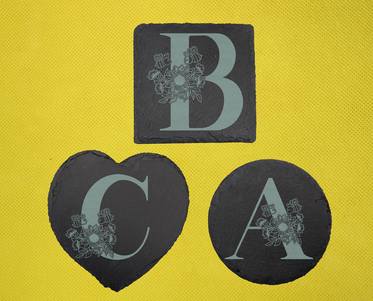 Shows 3 floral themed slate coasters, a square, heart and circle, engraved with the example initials A, B and C that have a sunflower, leaves and bees on and around them. It is on against a yellow background.