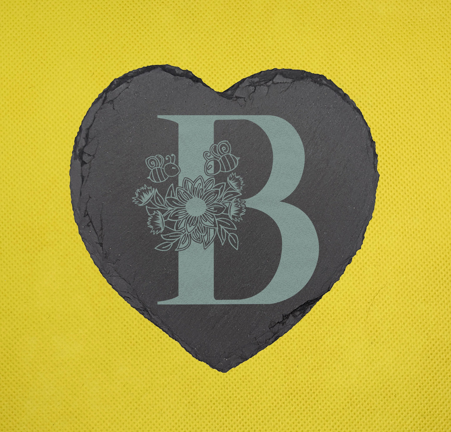 Shows a floral themed slate heart coaster engraved with the example initial B that has a sunflower, leaves and bees on and around it. It is against a yellow background.
