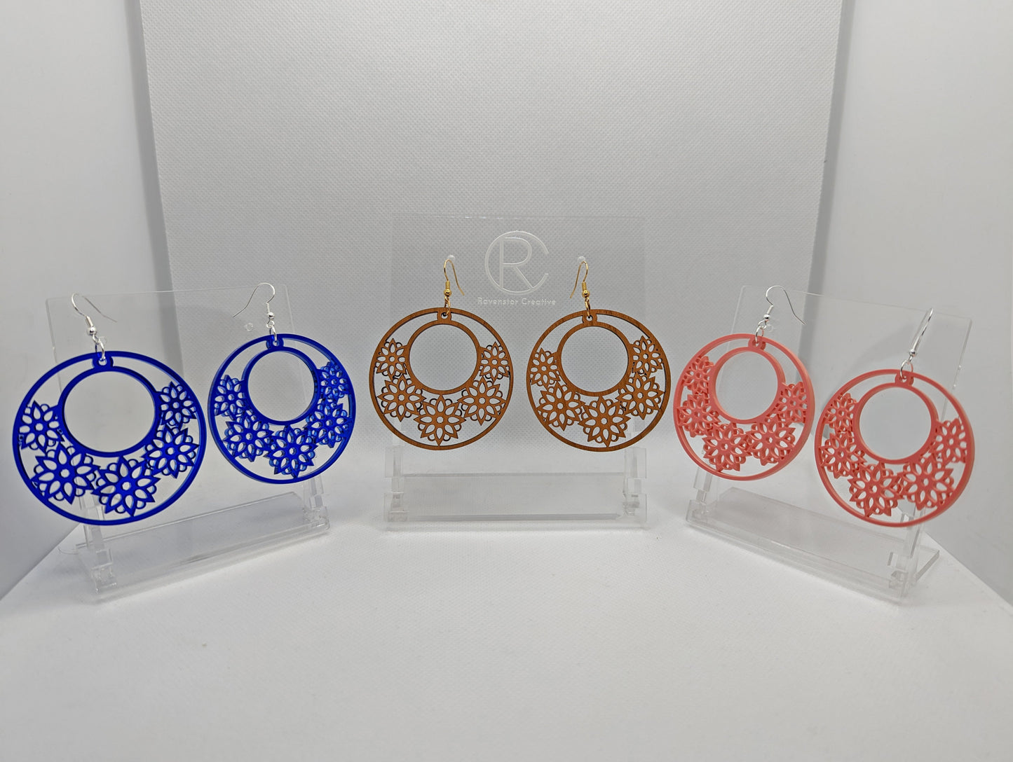 Three pairs of earrings with Ear wire fixings. They are large hoop earrings with flower / floral patterns cut in it. They are made from translucent blue acrylic, cherry wood and sherbet pink acrylic. They are on clear stands against white background.