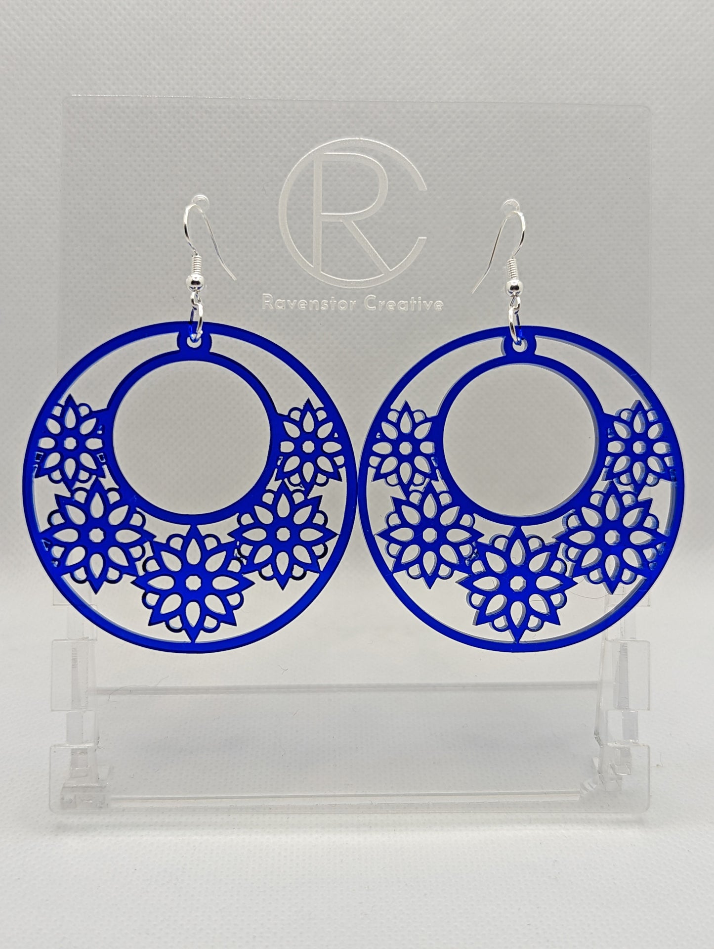 A pair of earrings with Ear wire fixings. They are large hoop earrings with flower / floral patterns cut in it. They are made from Translucent Blue acrylic. They are against a white background on a clear stand with the Ravenstor Creative logo on.