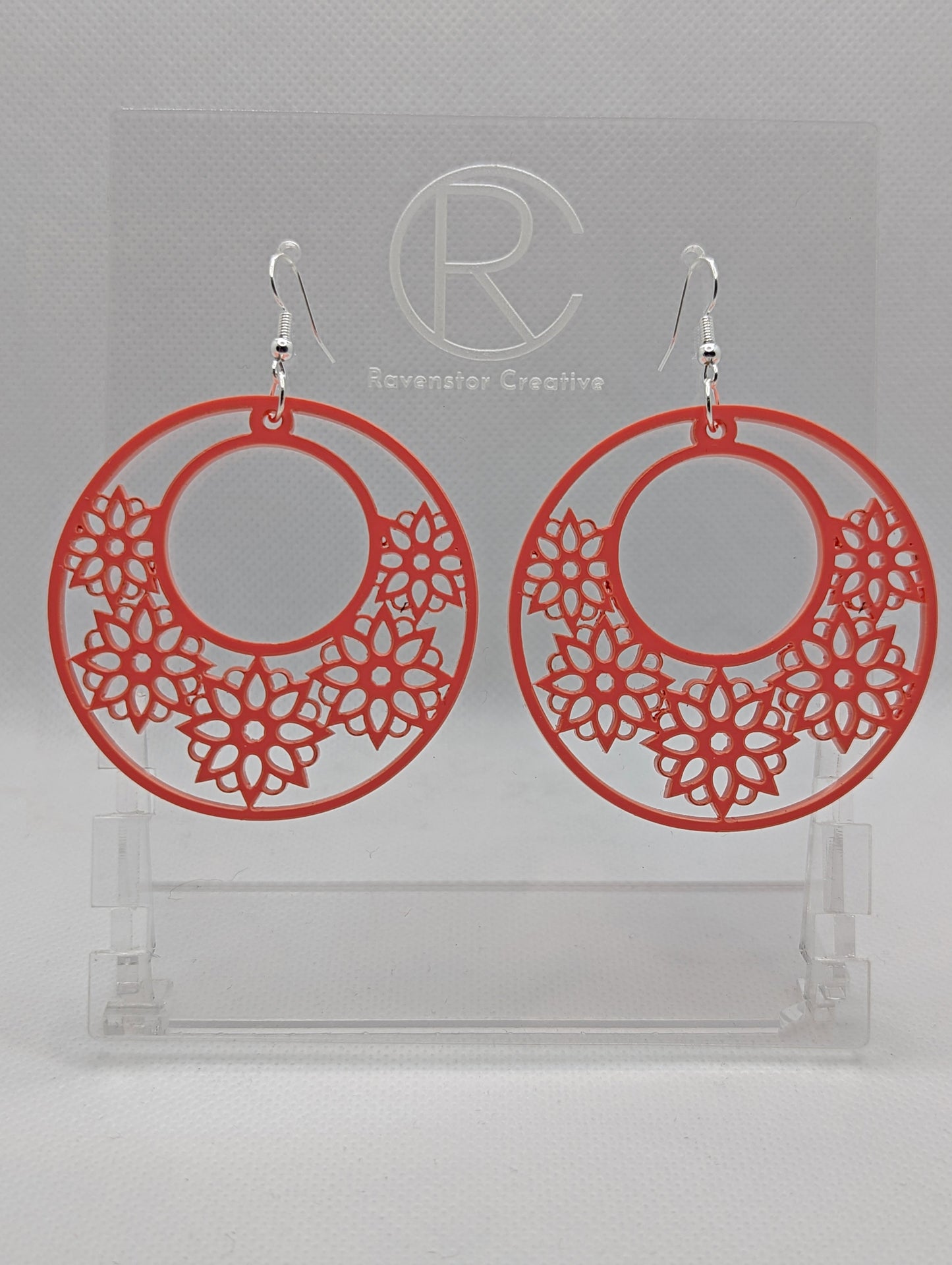 A pair of earrings with silver Ear wire fixings. They are large hoop earrings with flower / floral patterns cut in it. They are made from Sherbet Pink acrylic. They are against a white background on a clear stand with the Ravenstor Creative logo on.