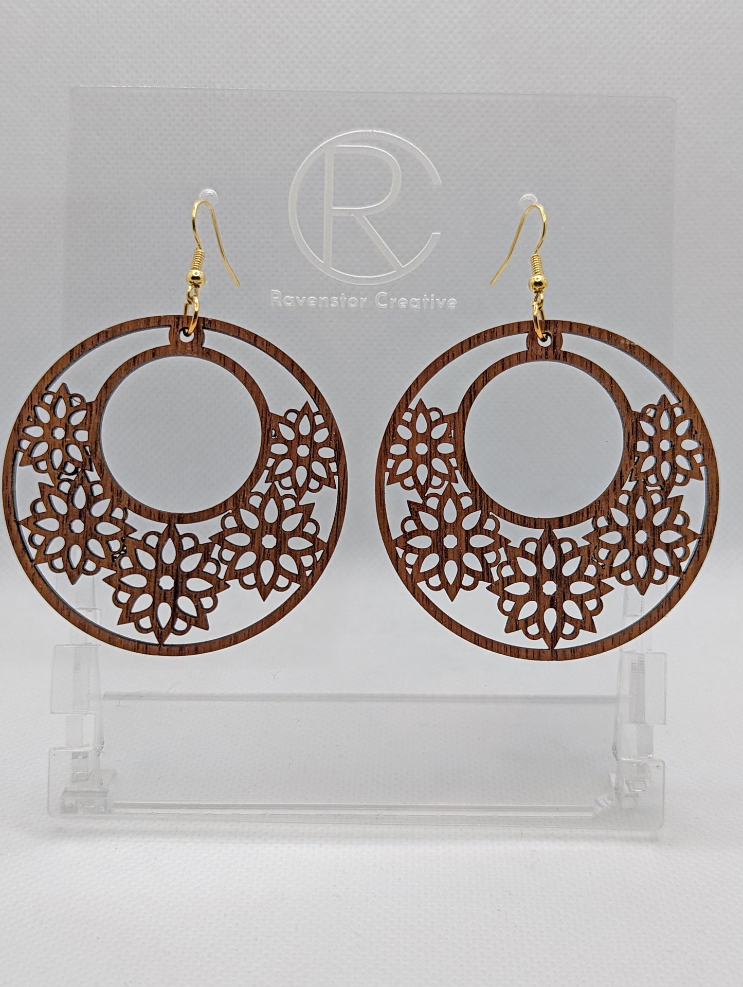 A pair of earrings with gold Ear wire hook fixings. They are large hoop earrings with flower / floral patterns cut in it. They are made from Walnut Wood. They are against a white background on a clear stand with the Ravenstor Creative logo on.