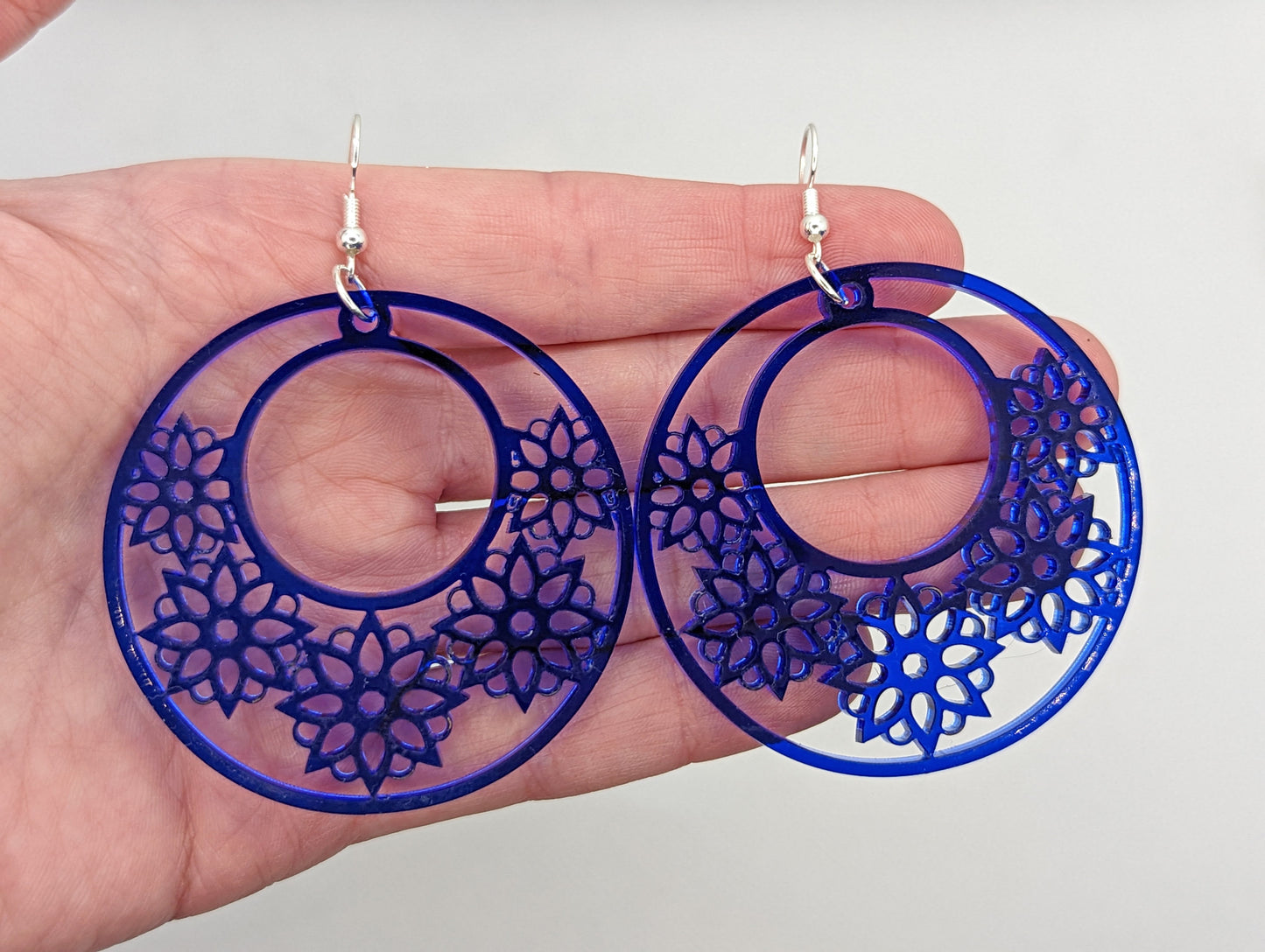 A pair of acrylic earrings against a hand showing the size. They are large hoop earrings with flower / floral patterns cut in it with silver ear wire hook fixings. They are 4 finger widths in length and same again wide. The colour is translucent blue