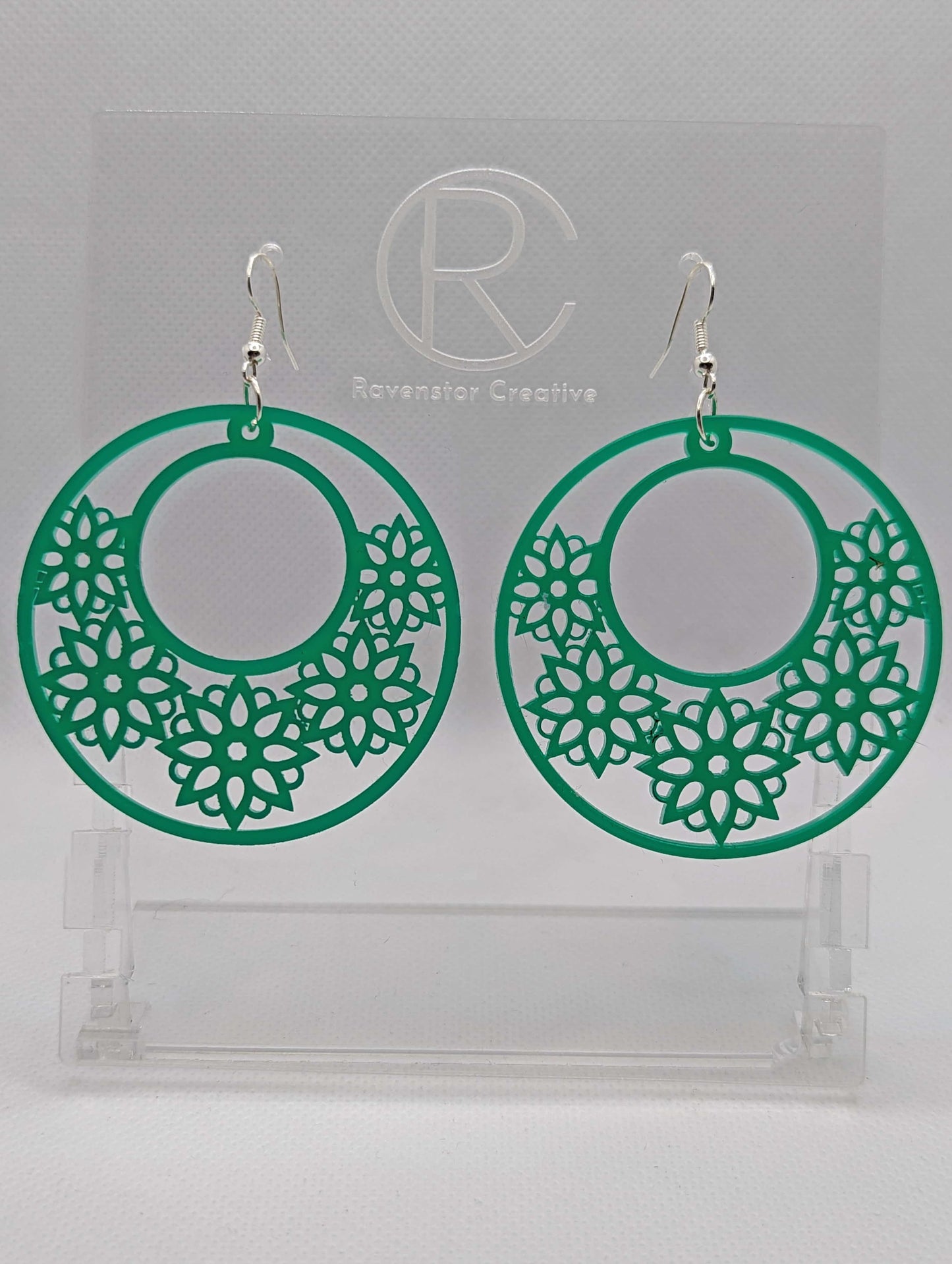 A pair of earrings with silver Ear wire hook fixings. They are large hoop earrings with flower / floral patterns cut in it. They are made from Green acrylic. They are against a white background on a clear stand with the Ravenstor Creative logo on.