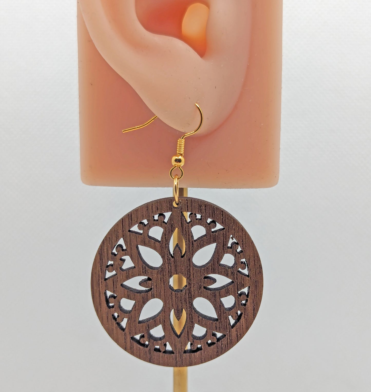 Shows a Floral Themed Circle earring with an intricate pattern cut out of the middle, its on an ear showing the size against a white background. it is Walnut wood and has Gold fixings.