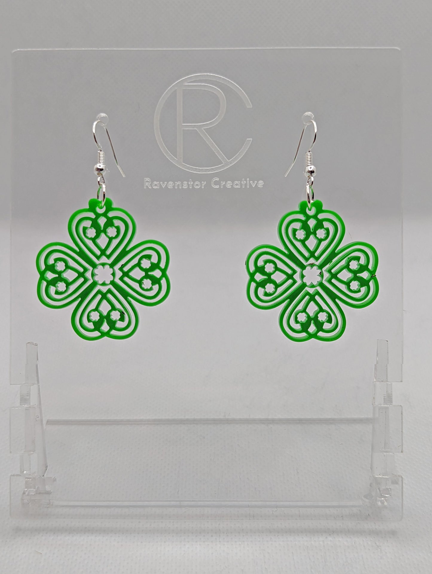 Shows a pair of acrylic earrings that have silver ear wire hook fixings. They the a four leaf clover design with a Celtic pattern. They are Green in colour and are against a white background on see through stand with the Ravenstor Creative logo on it