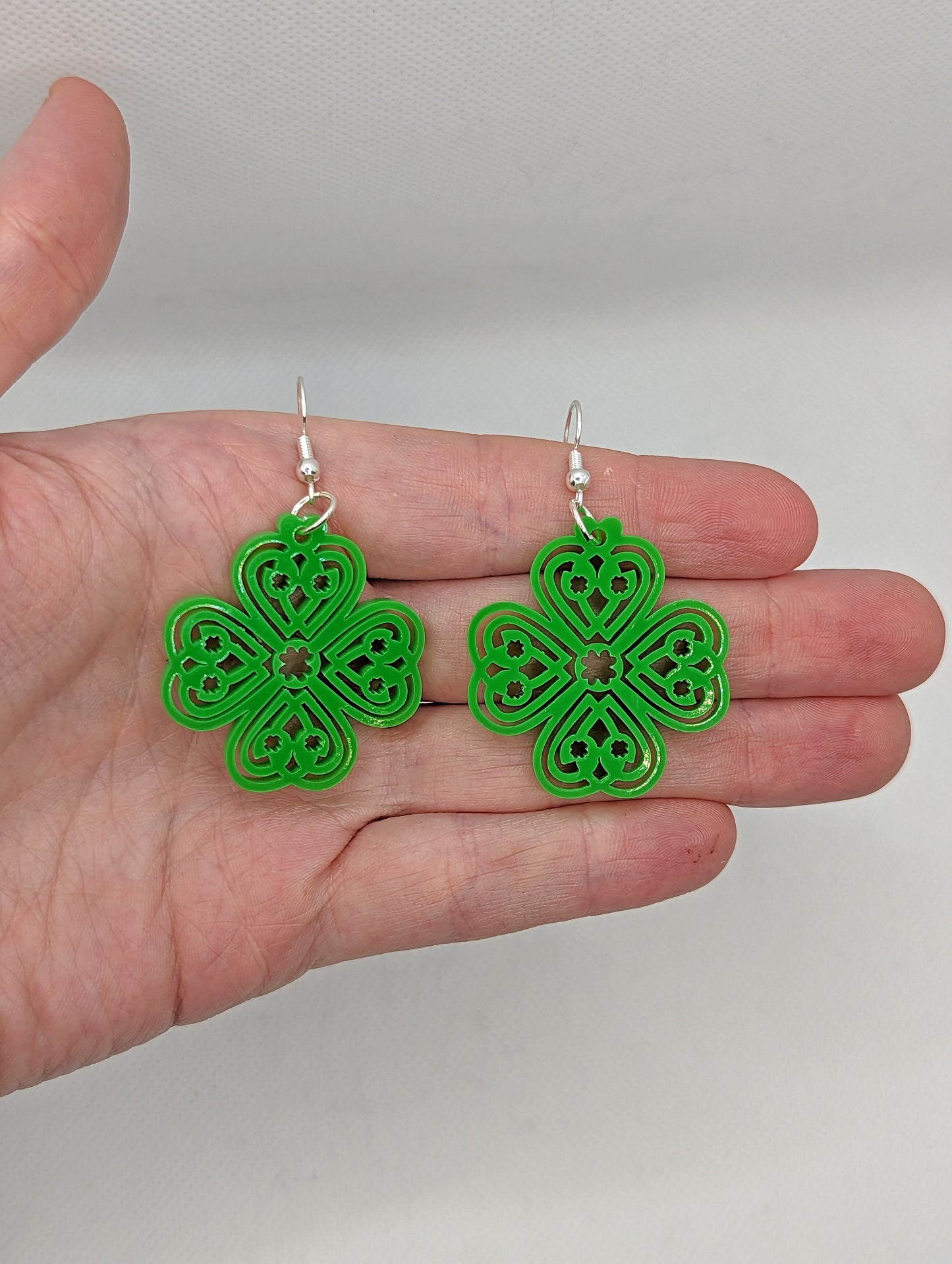 Shows a pair of acrylic earrings that have silver ear wire hook fixings. They are four leaf clover design with Celtic patterns. They are against a hand showing the size of two and a half finger widths in length and same again wide.