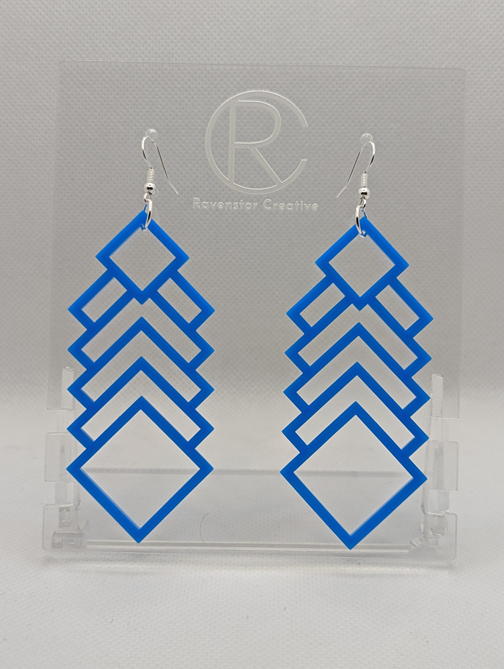 A pair of large and long acrylic geometric diamond themed earrings. They have silver ear wire hook fixings and they are Blue in colour. They are on a clear stand against a white background that has the Ravenstor Creative logo on it.