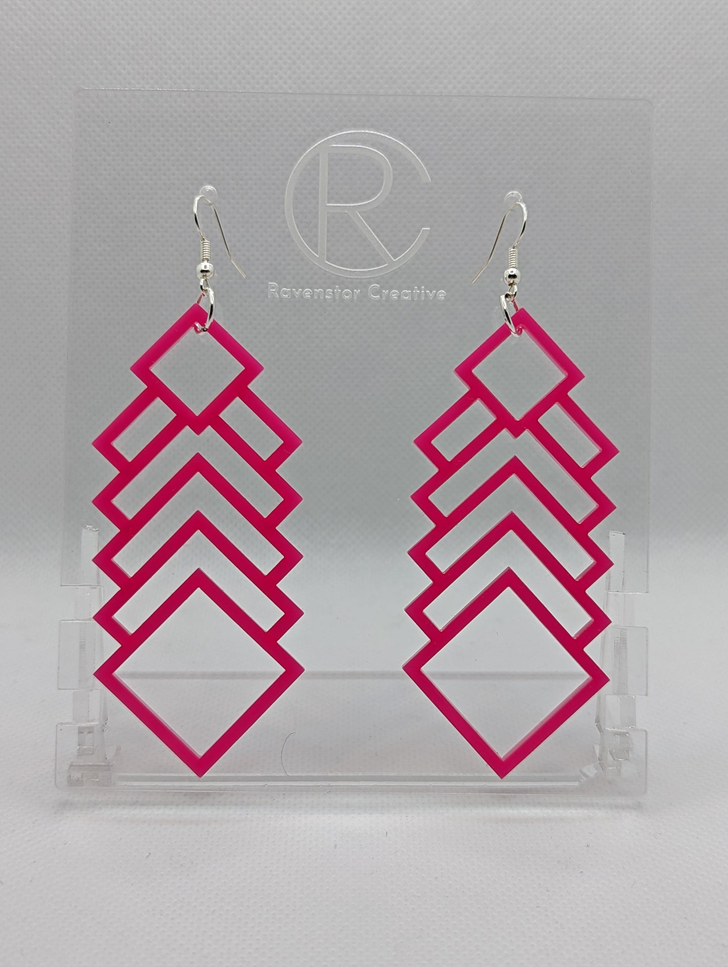 A pair of large and long acrylic geometric diamond themed earrings. They have silver ear wire hook fixings and they are Hot Pink in colour. They are on a clear stand against a white background that has the Ravenstor Creative logo on it.