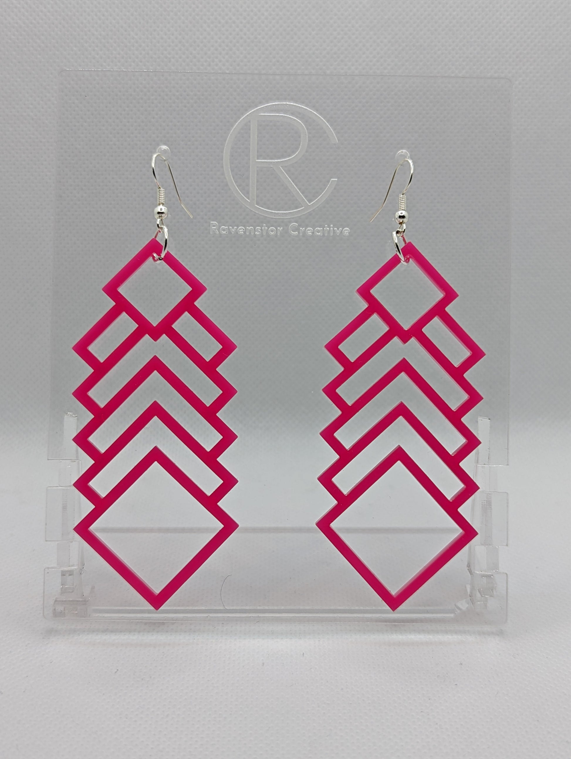 A pair of large and long acrylic geometric diamond themed earrings. They have silver ear wire hook fixings and they are Hot Pink in colour. They are on a clear stand against a white background that has the Ravenstor Creative logo on it.