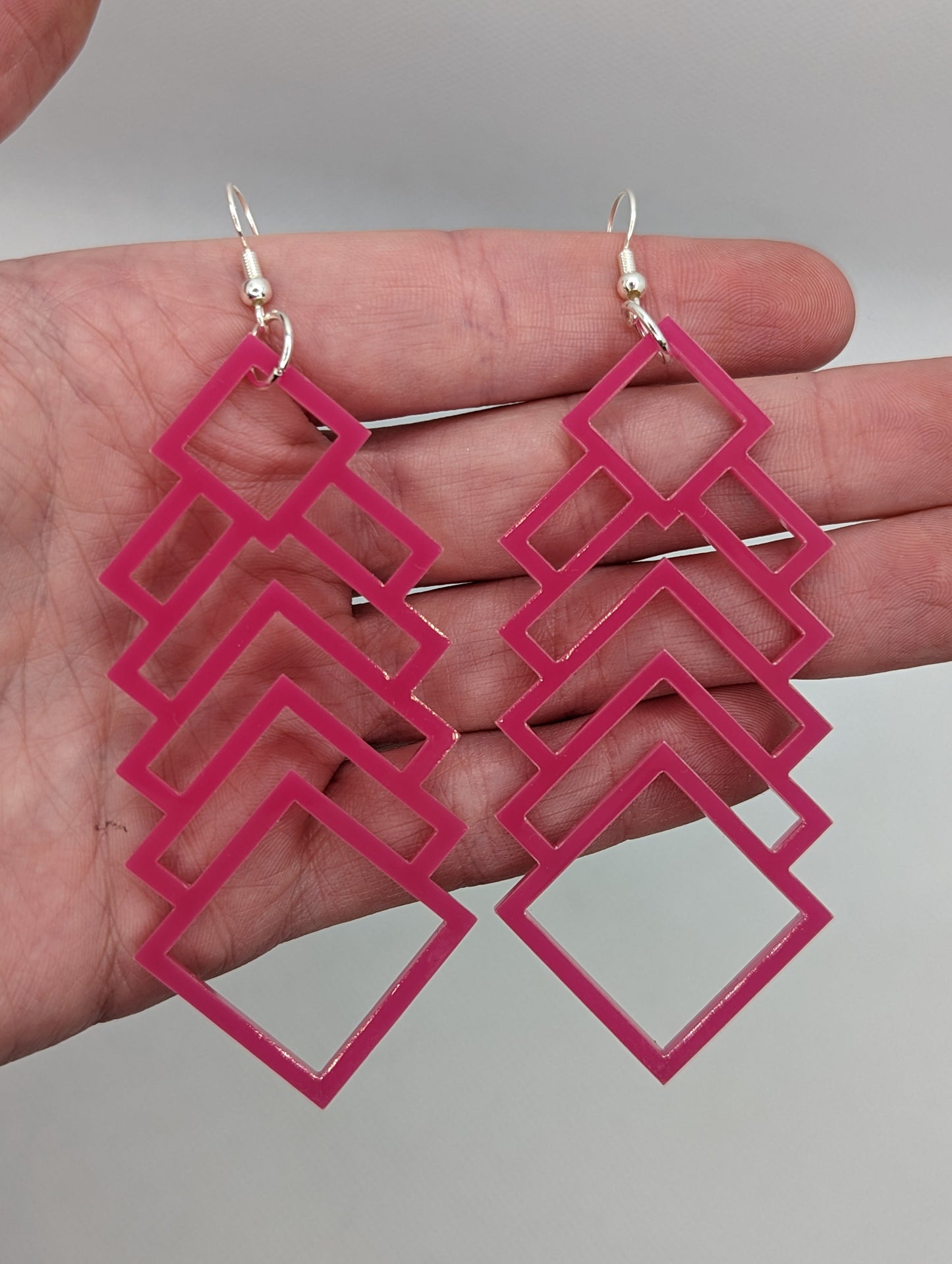 A pair of large and long acrylic geometric diamond themed earrings. They have ear wire hook fixings and are against a hand to show the size.  They are five finger widths in length and two and a half wide.