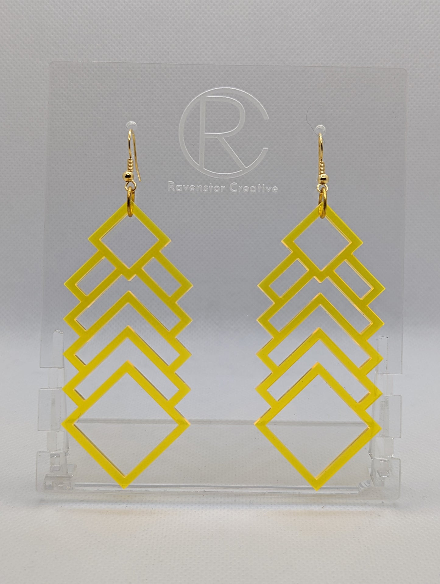A pair of large and long acrylic geometric diamond themed earrings. They have Gold ear wire hook fixings and they are Neon Yellow in colour. They are on a clear stand against a white background that has the Ravenstor Creative logo on it.
