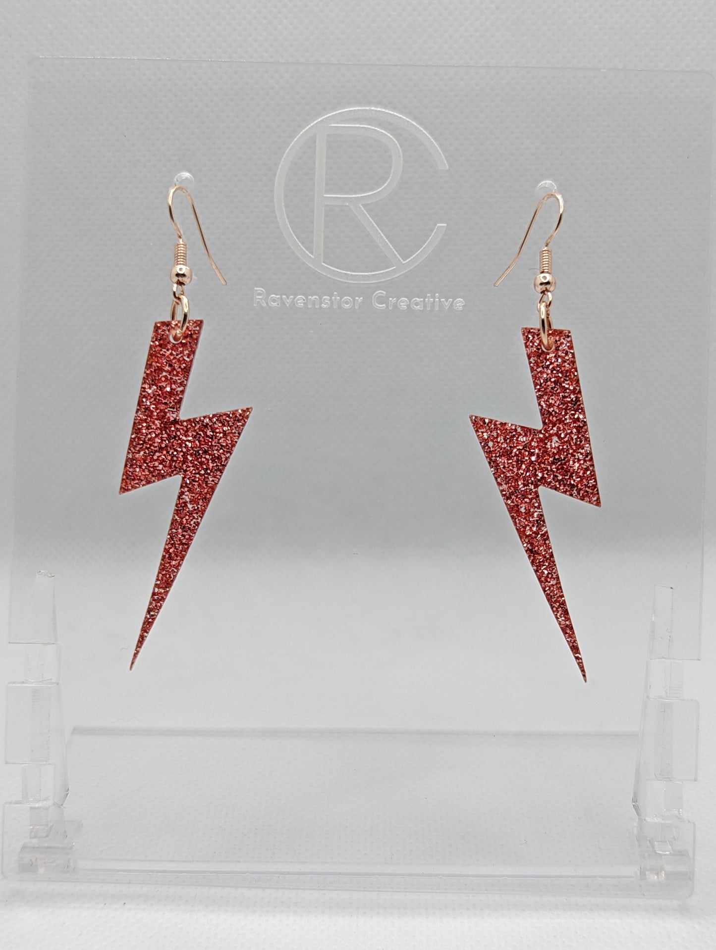 Shows a pair of earrings that have Rose Gold ear wire hook fixings. They are Lightning Bolt shapes similar to Harry Potter's forehead. They are Copper Glitter colour and are against a white background on see through stand.