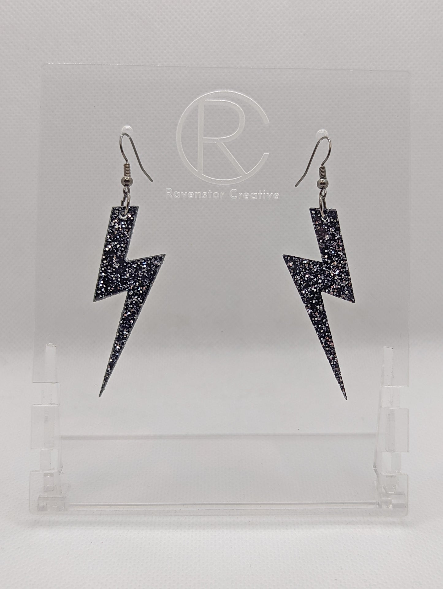 Shows a pair of earrings that have black ear wire hook fixings. They are Lightning Bolt shapes similar to Harry Potter's forehead. They are Black & Silver Glitter colour and are against a white background on see through stand.