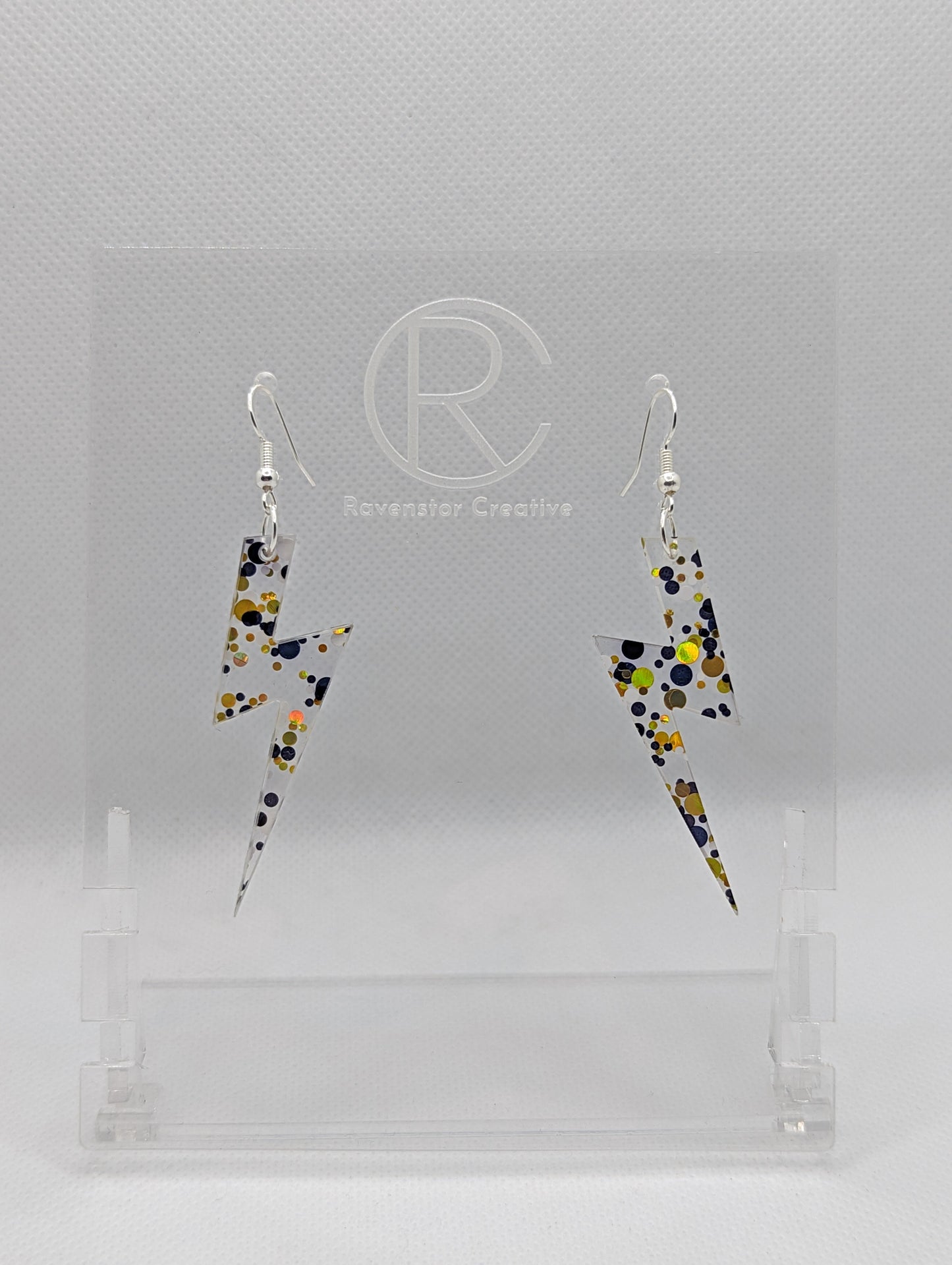 Shows a pair of earrings that have silver ear wire hook fixings. They are Lightning Bolt shapes similar to Harry Potter's forehead. They are Lollipop Glitter colour and are against a white background on see through stand.