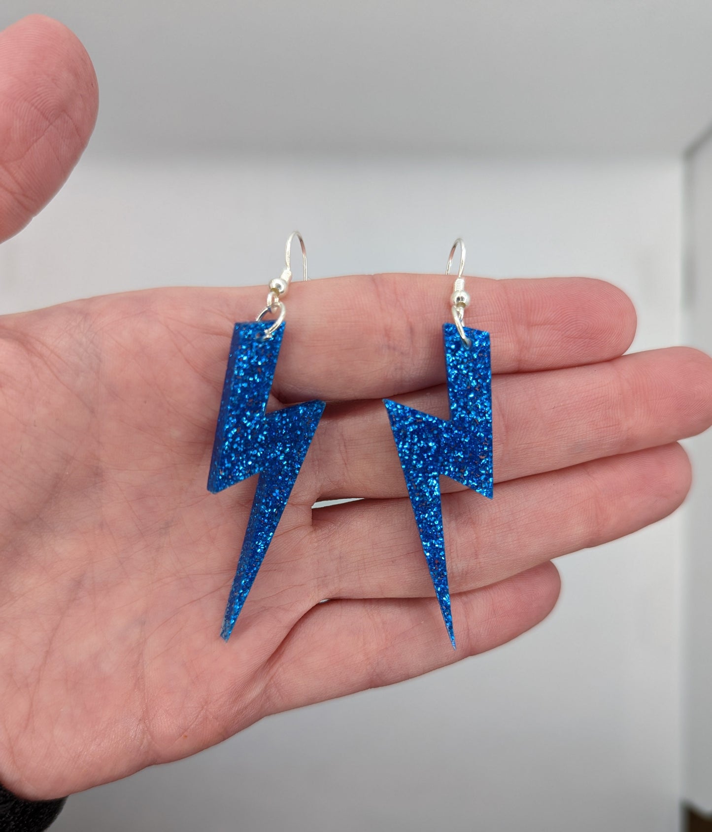 Image of a pair of lightning bolt shaped earrings on a hand showing the size against a white background, they are three and a half finger widths length and one and a half wide. They are royal blue glitter in colour and have silver fixings.